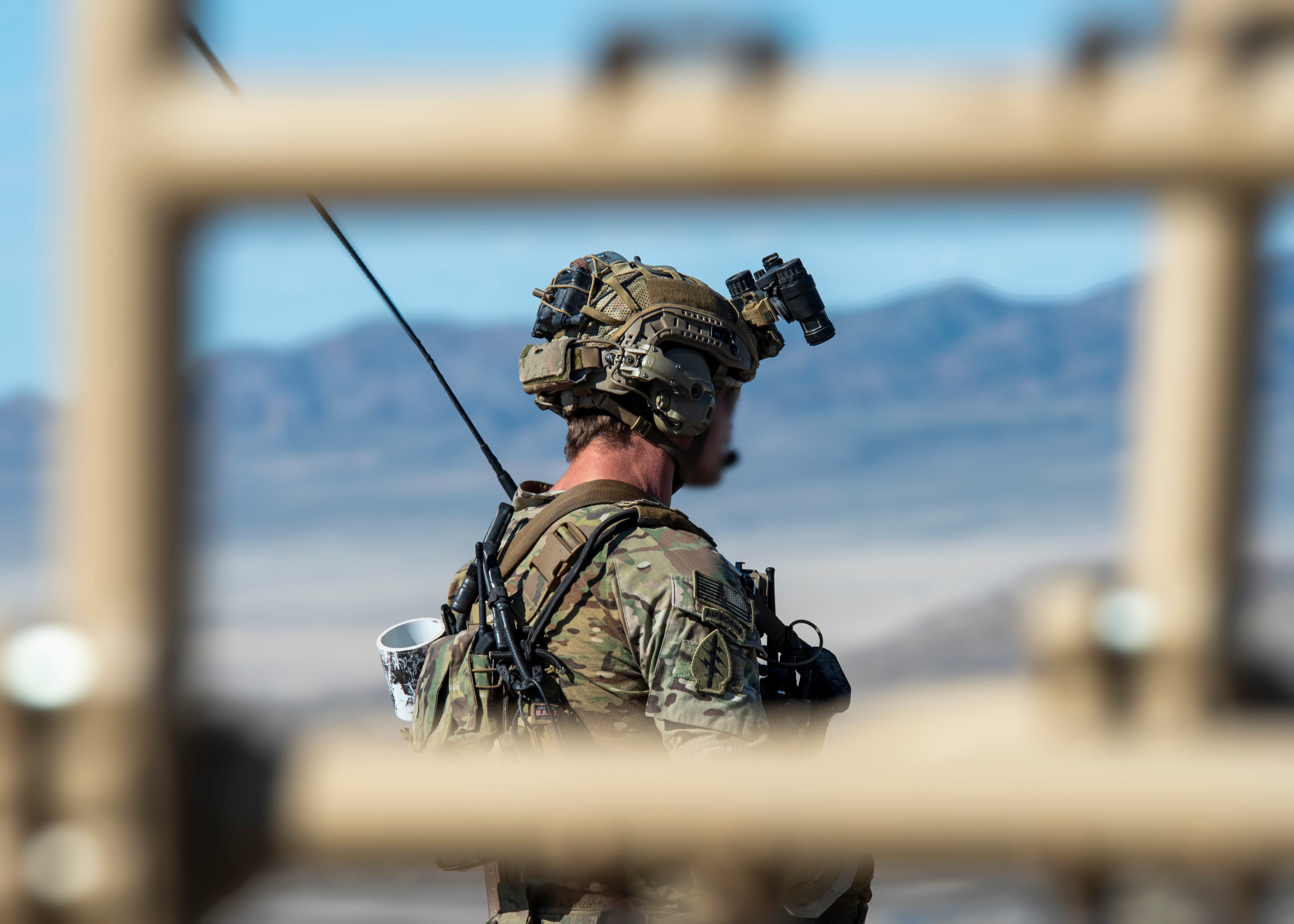 The right-side and back of a man wearing camouflage and a helmet with a scope on it are seen through blurred out tope-colored bars with a distant mountain landscape in the background. The man's face is blurred out.