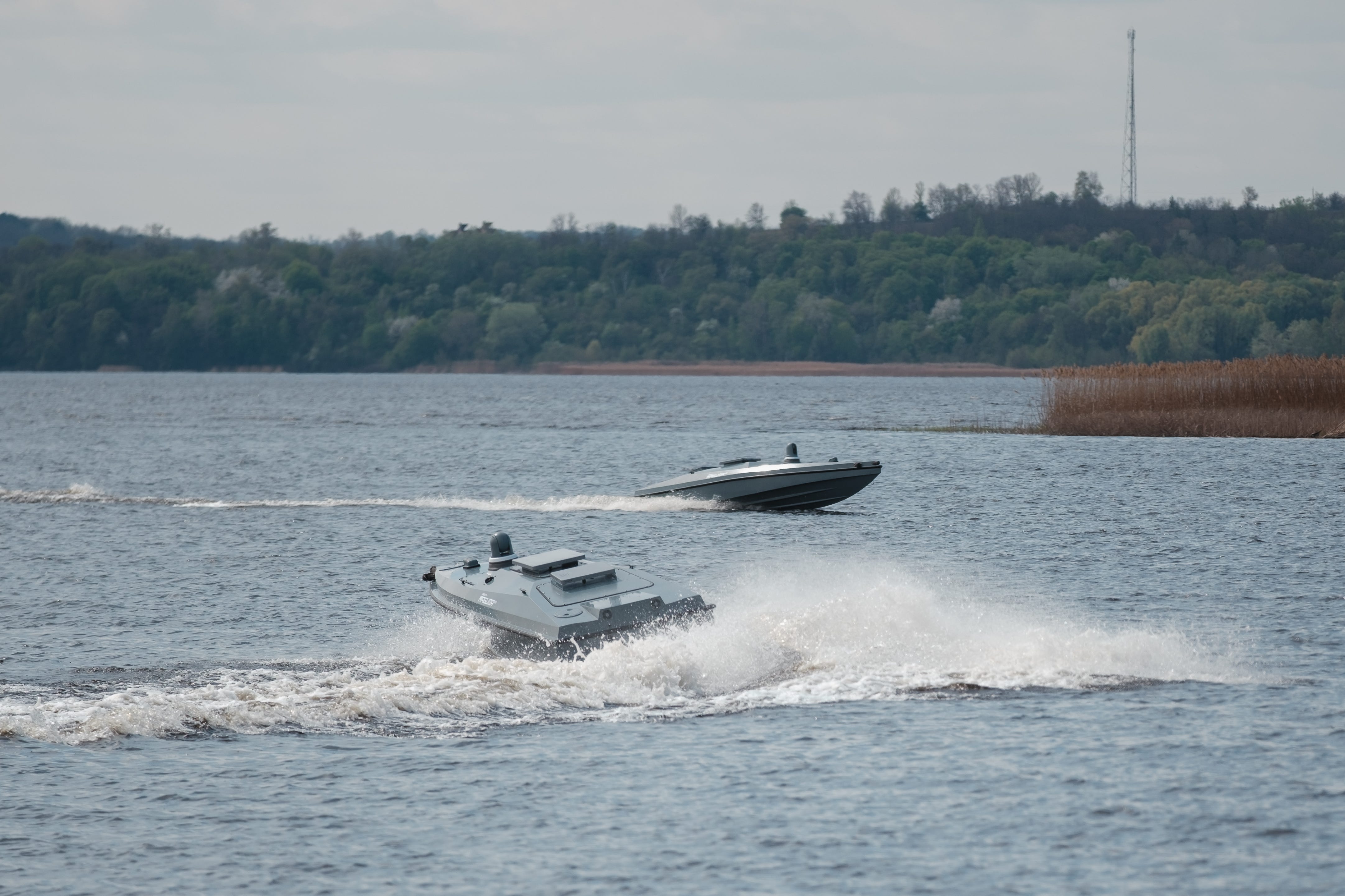 Two grey naval drones move in a body of water with trees behind them