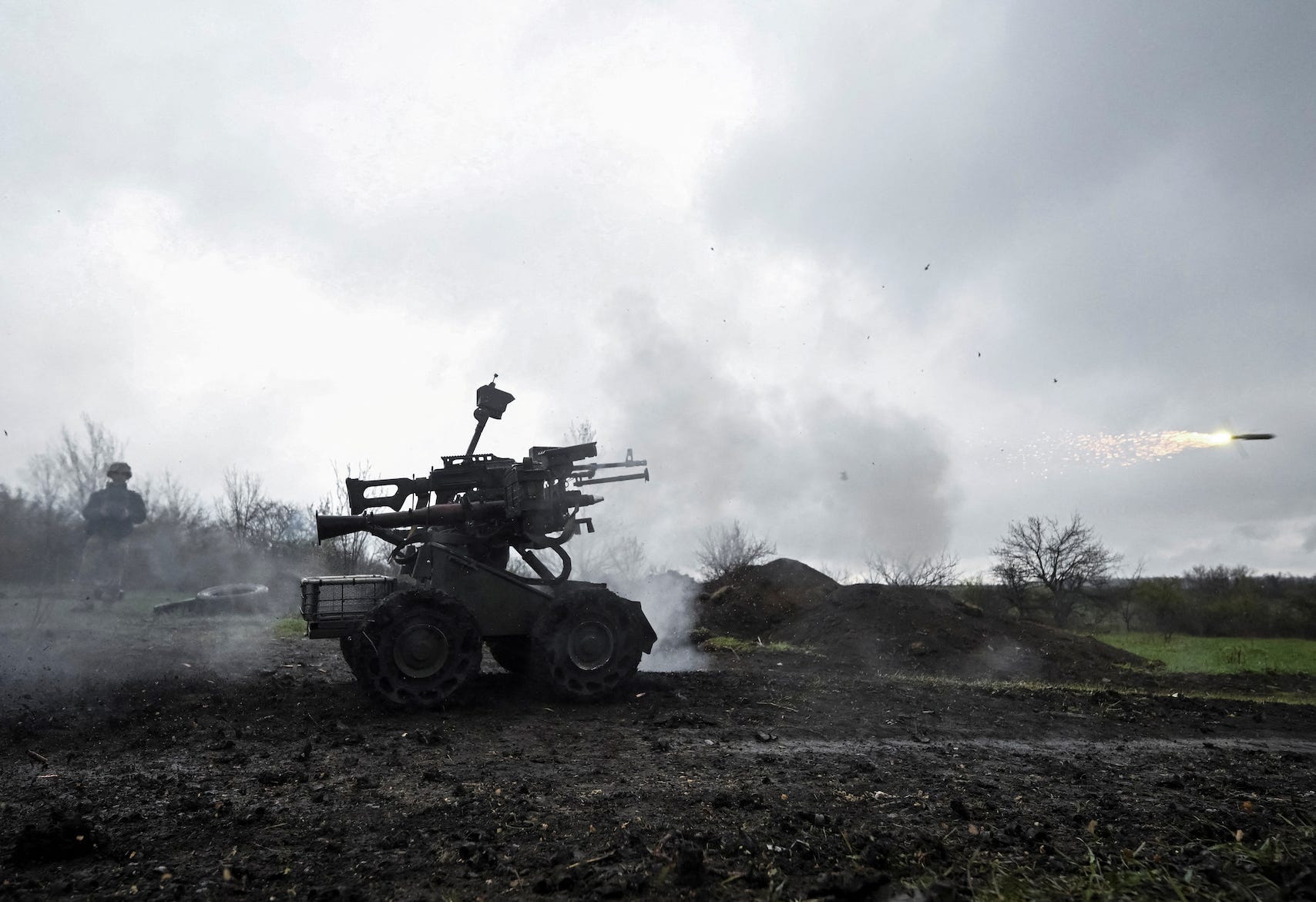 A small wheeled robot fitted with a grenade launcher rolls on a dirt ground with a grey sky and a man behind it