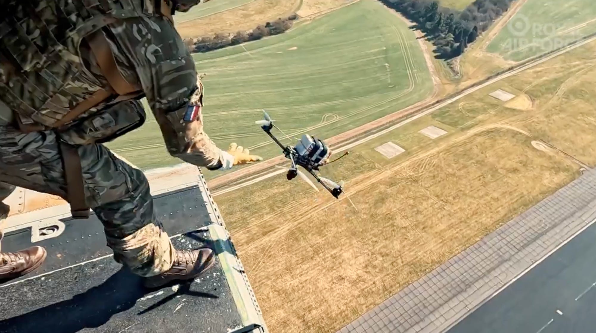 A still from footage taken inside a helicopter shows a figure in camouflage gear letting go of a small drone with a brown field and a runway below