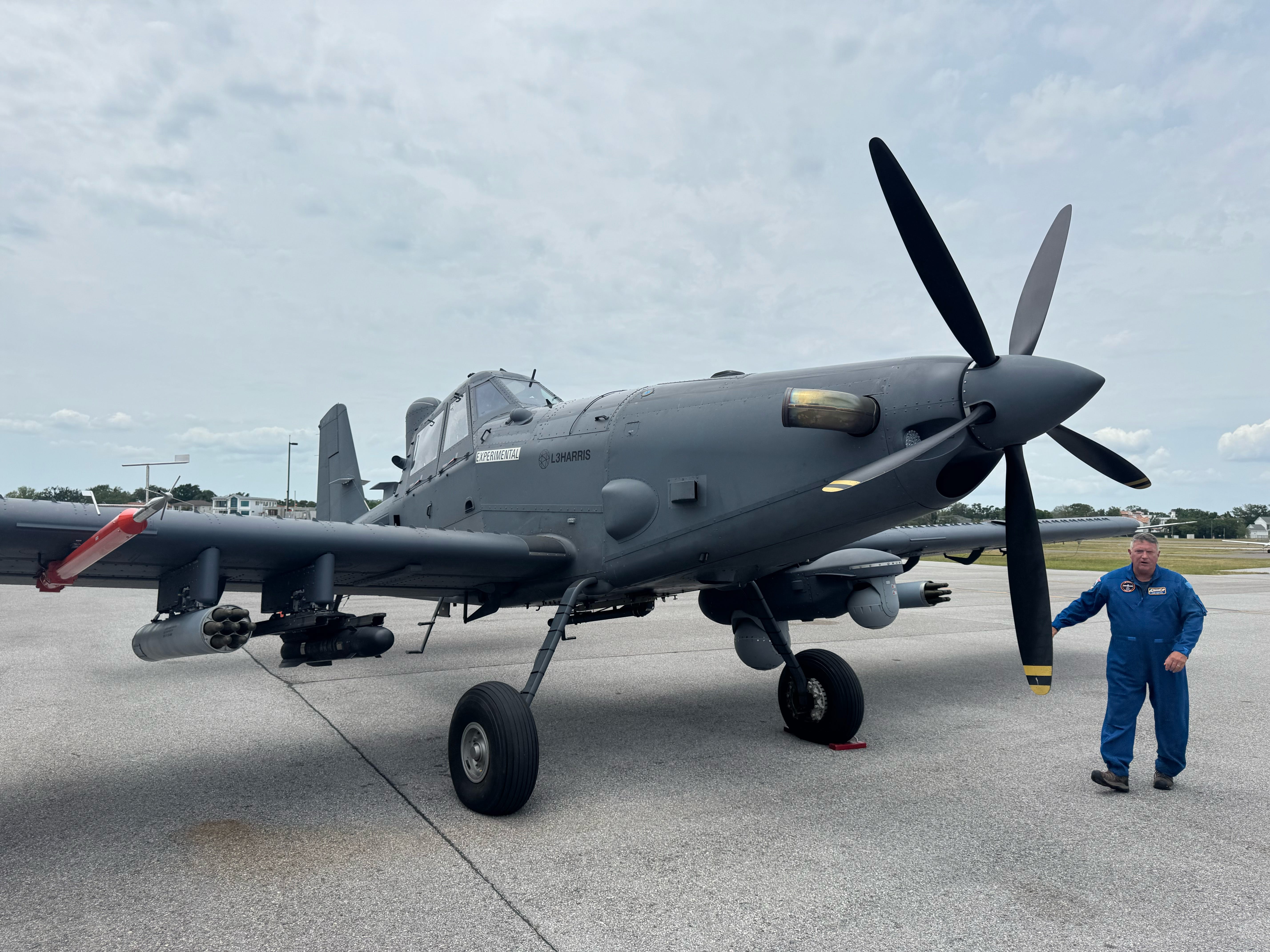 L3Harris' Sky Warden airplane, grey in color, sitting on an airfield with a man wearing a blue flight test suit standing next to it. The sky is overcast in the background.