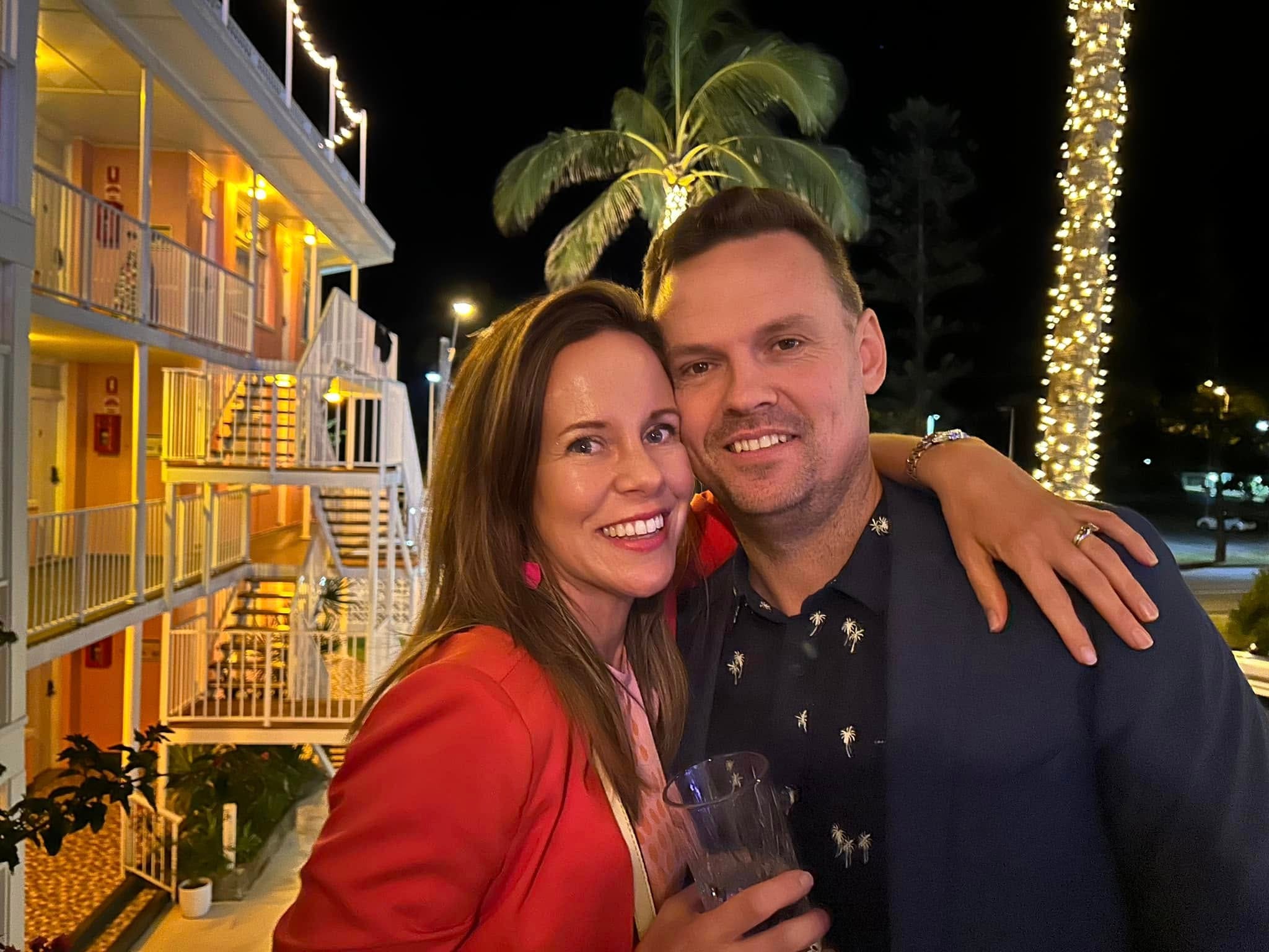 The author and her husband standing outside on a hotel balcony at night with a palm tree and lights outside.