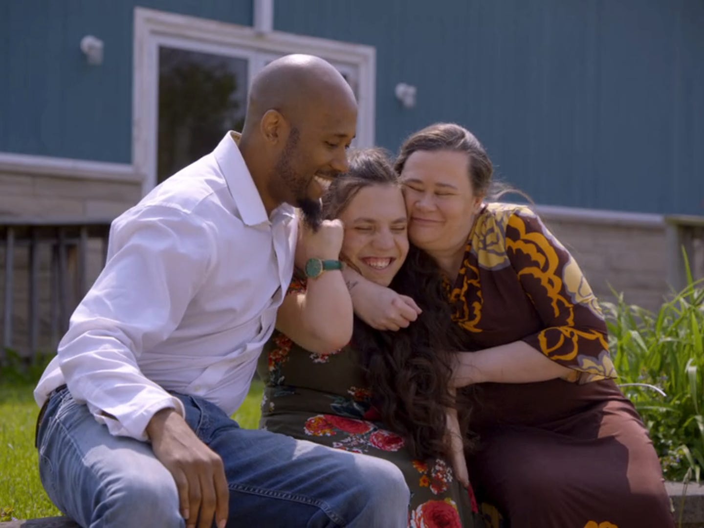 antwon mans, natalia grace mans, and cynthia mans sitting together outside a blue building. they're all smiling and hugging each other tightly, with natalia sitting in the middle