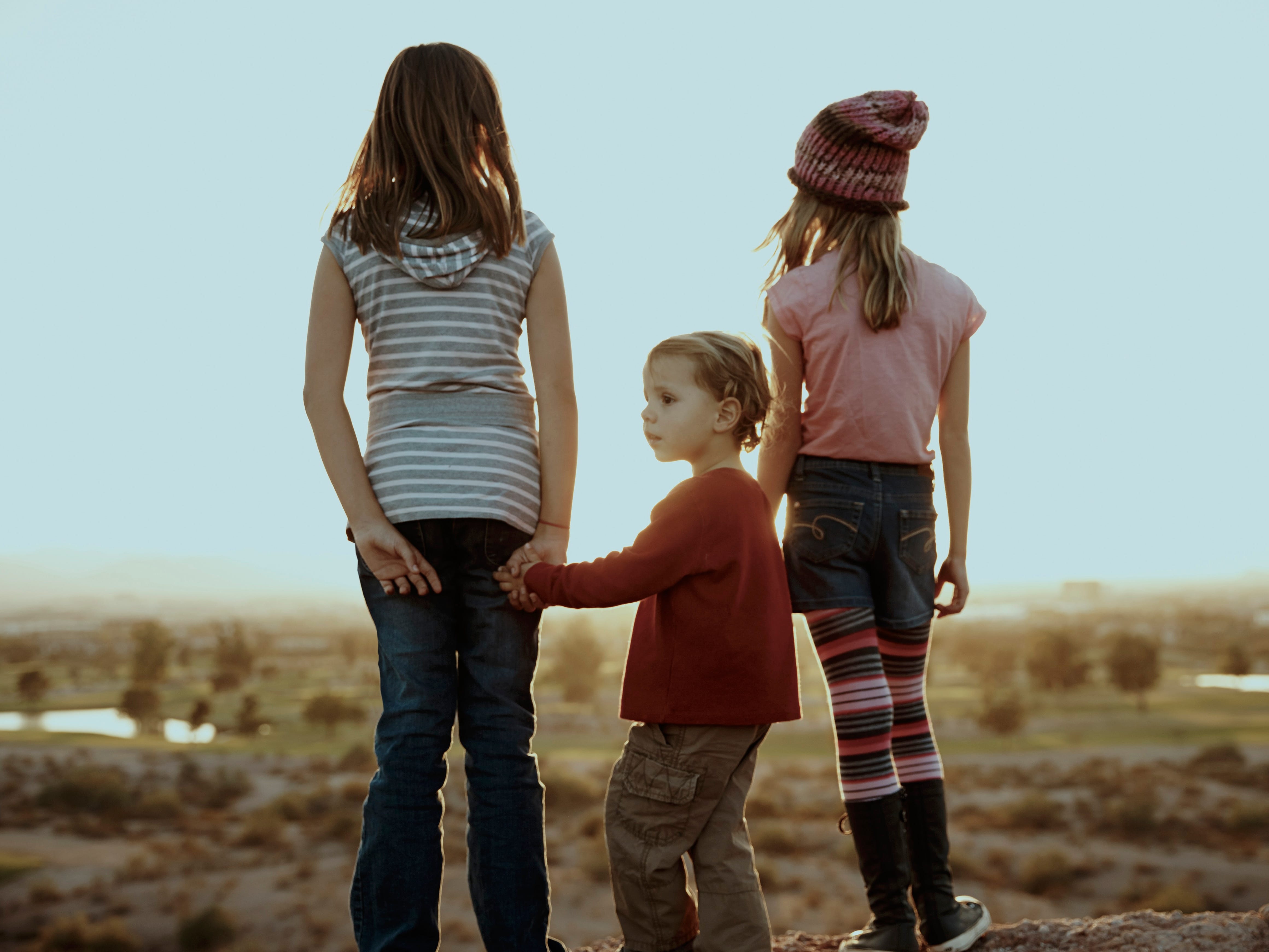Three kids standing and looking at the sunset holding hands.
