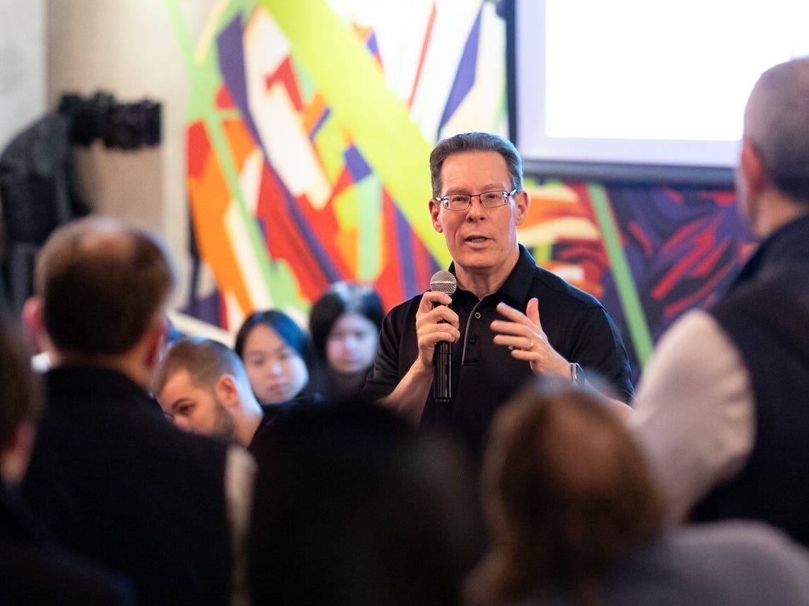 A man wearing glasses is holding a microphone and speaking to a crowd.