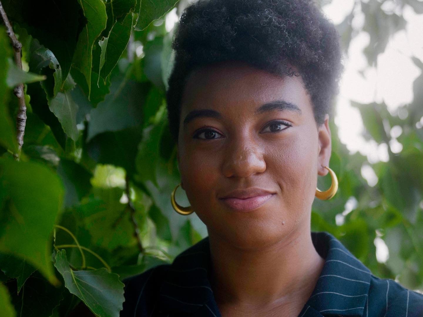 Natalie Jasmine Harris wearing a pinstripe vest and standing in front of trees.