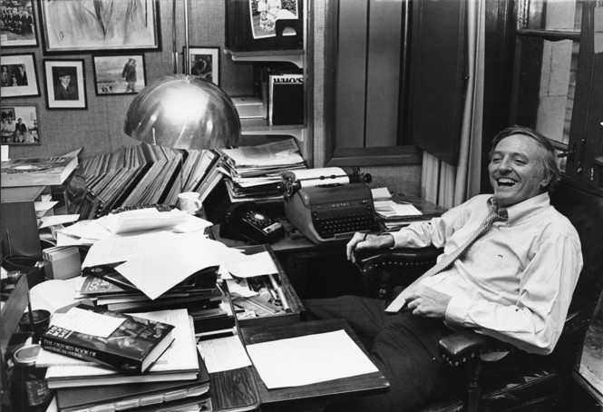 A black and white photo of William F. Buckley laughing while seated at his desk, papers and books all around him