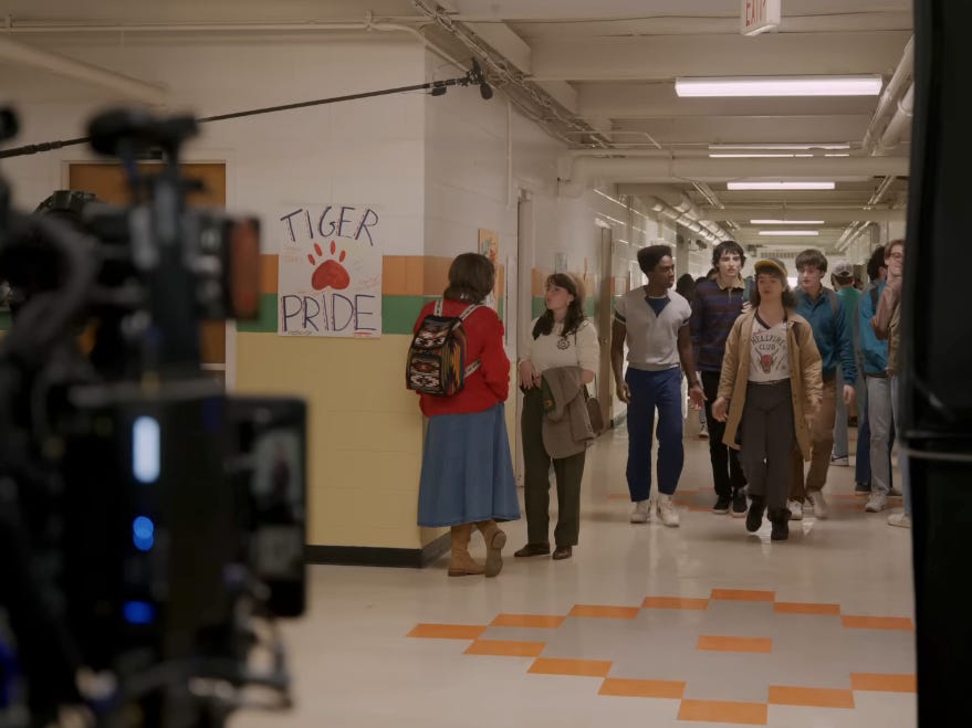the stranger things season five set. a high school hallway filled with students, and actors caleb mclaughlin, finn wolfhard, gaten matarazzo, and noah schnapp walking down as their characters lucas, mike, dustin, and will. in the foreground, you can see a camera and boom microphone