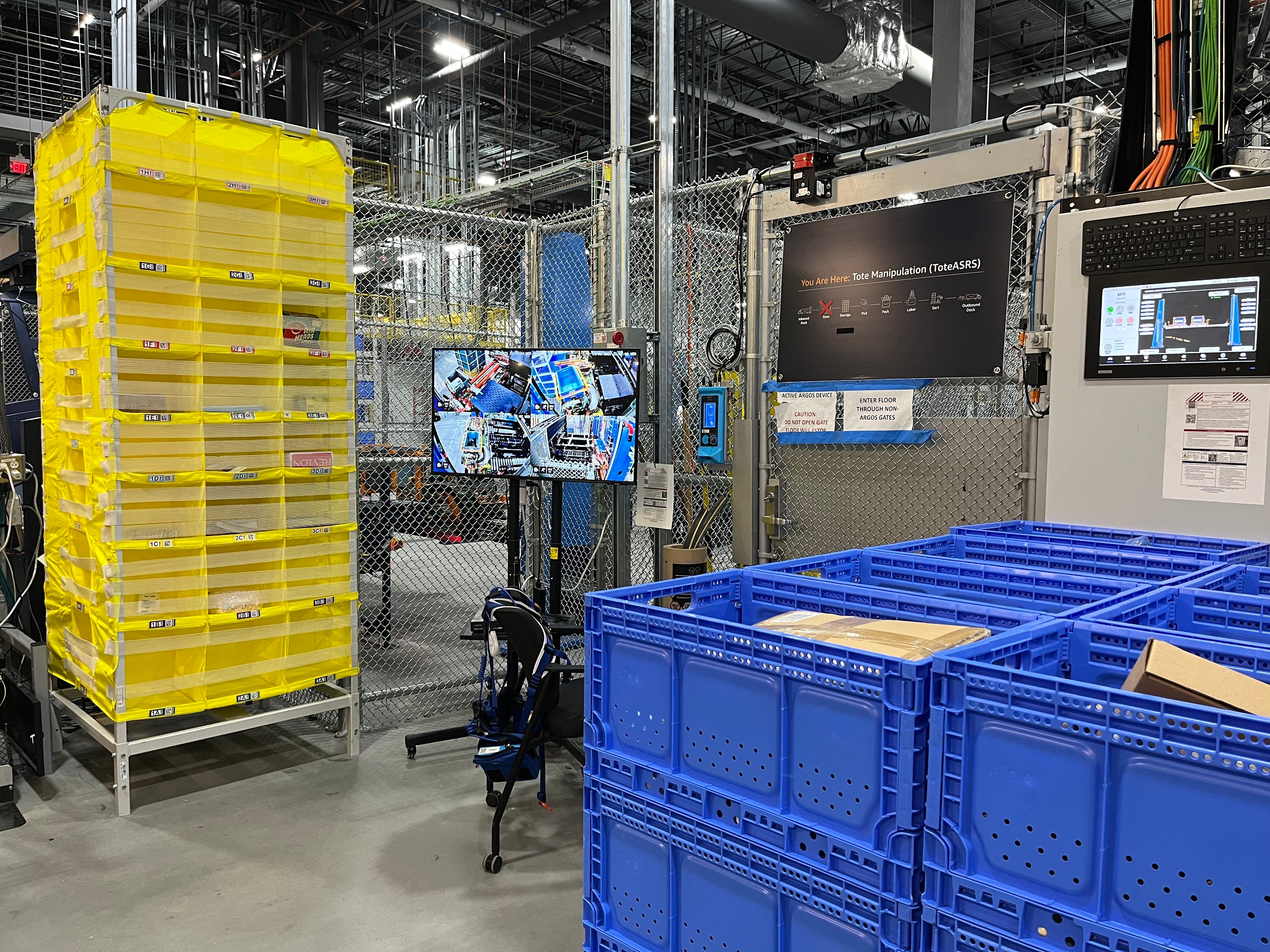 Yellow cart with products inside sits by blue bins and monitors inside Amazon Robotics