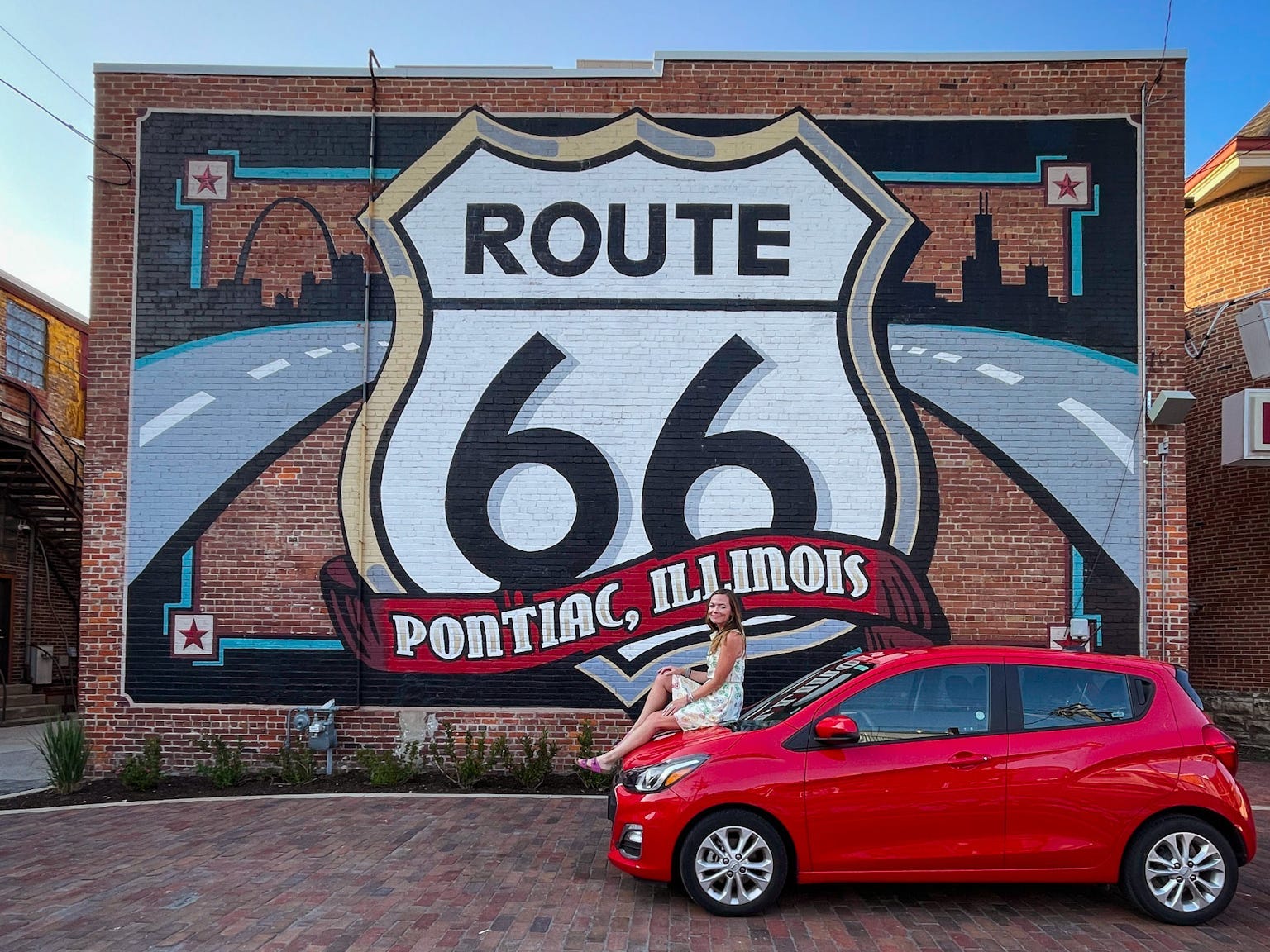 Emily sits on the hood of a red car in front of a mural that reads "Route 66 Pontiac, Illinois."