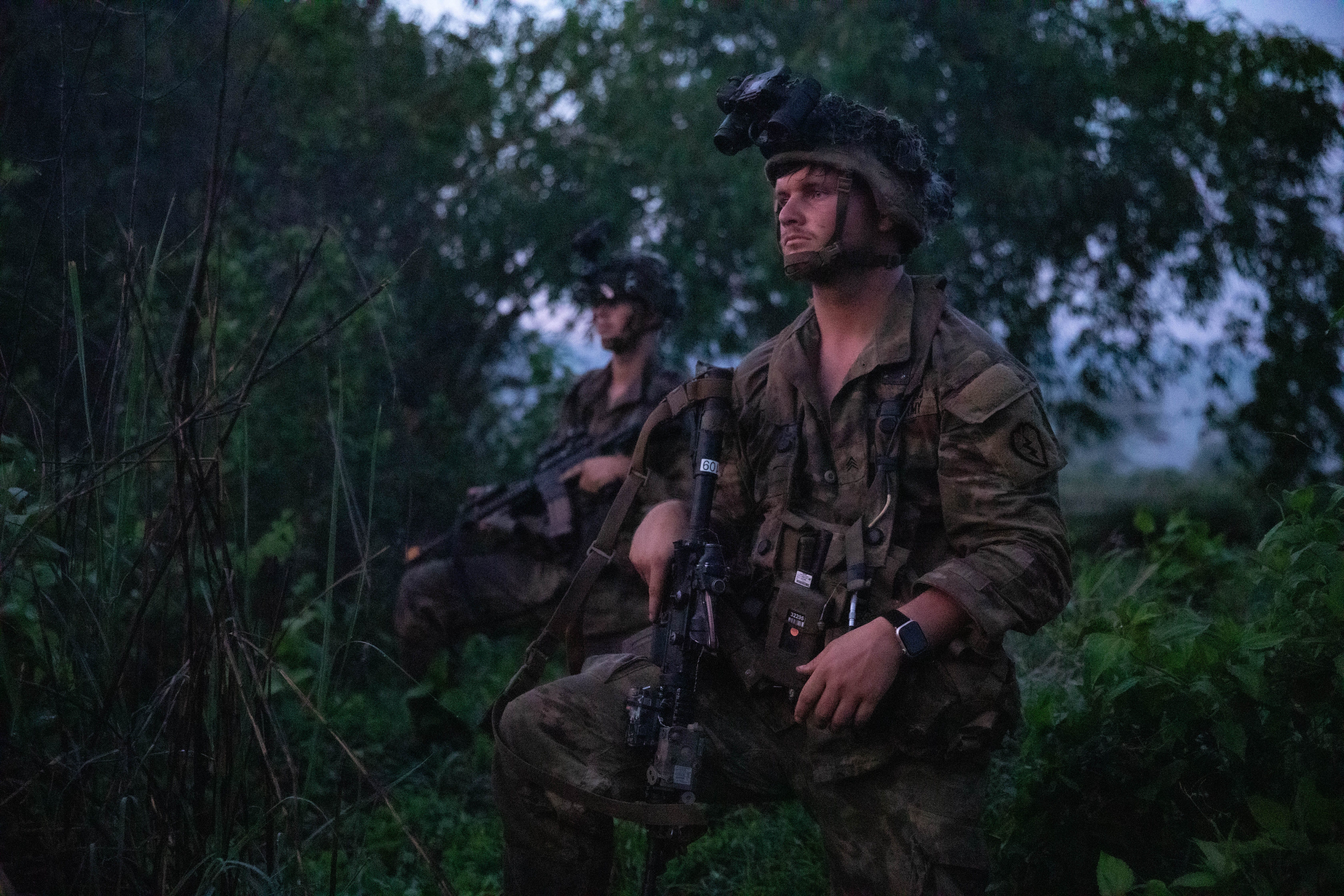 A man wearing camouflage kneels holding a rifle. Another man is doing the same thing in the background. The two men are surrounded by trees and grass.
