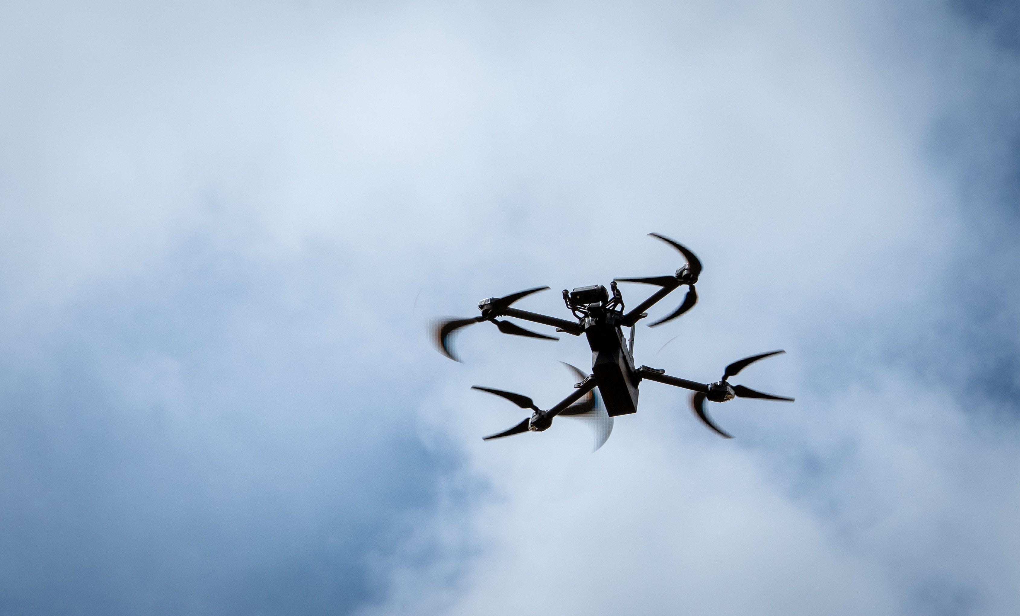 A small black quadcopter drone flies with a cloudy blue sky in the background.