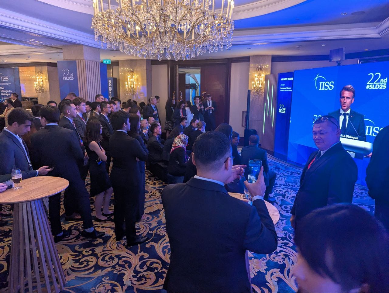 Staffers crowd a screen outside the Shangri-La Island Ballroom while watching a screen displaying French President Emmanuel Macron.