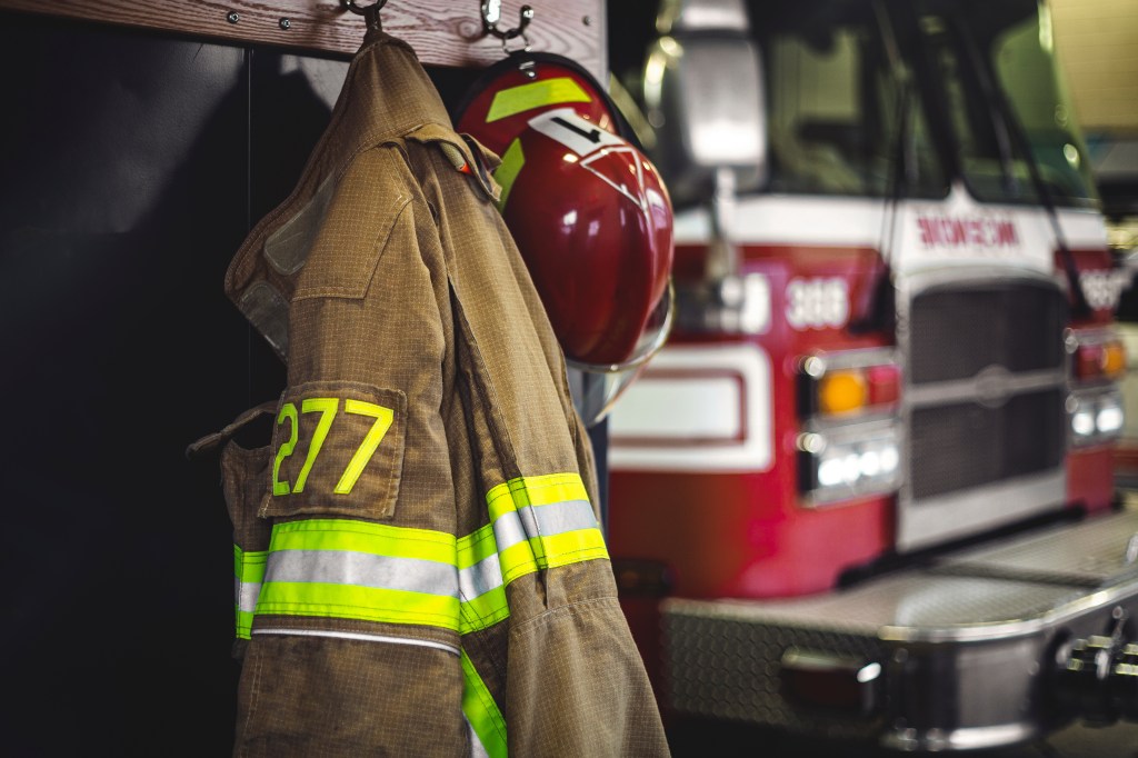 Firefighter jacket and helmet hanging in firehouse with firetruck in background.