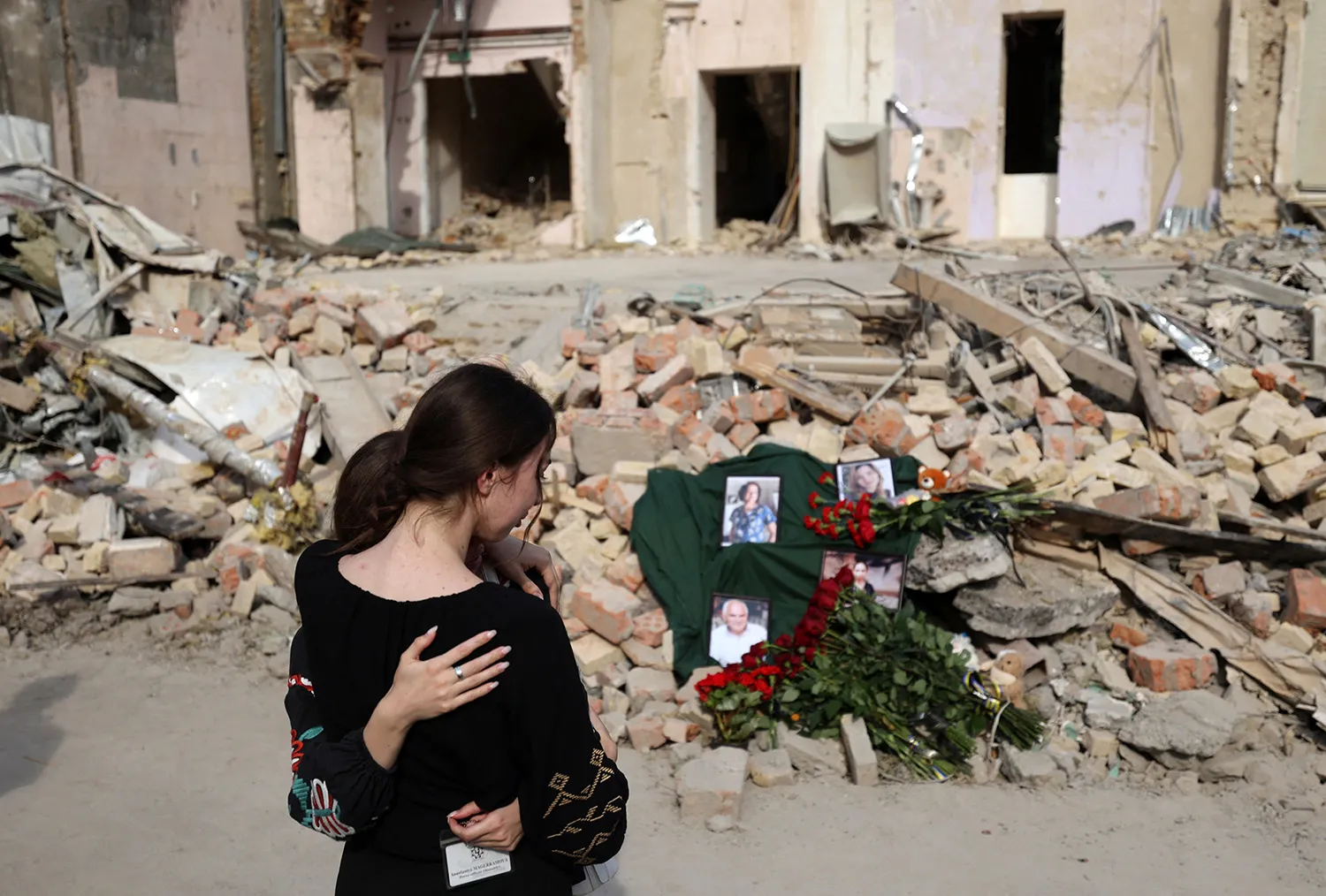 Two people embrace as they look down at flowers and photos atop a pile of rubble outside of a damaged building.