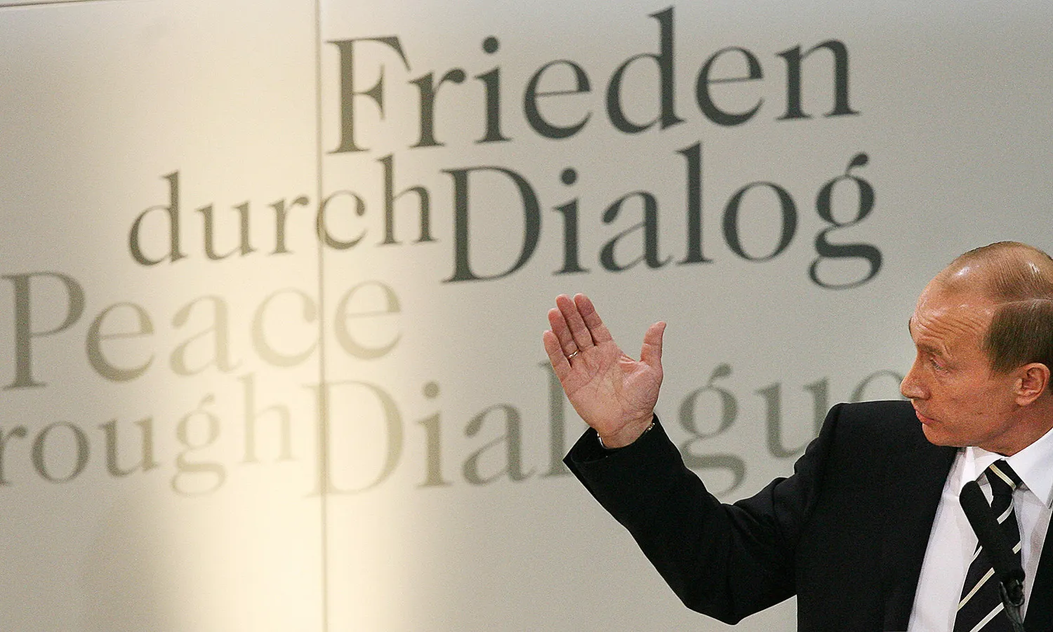 Vladimir Putin gestures with one hand as he stands in front of a wall with words in German on it.