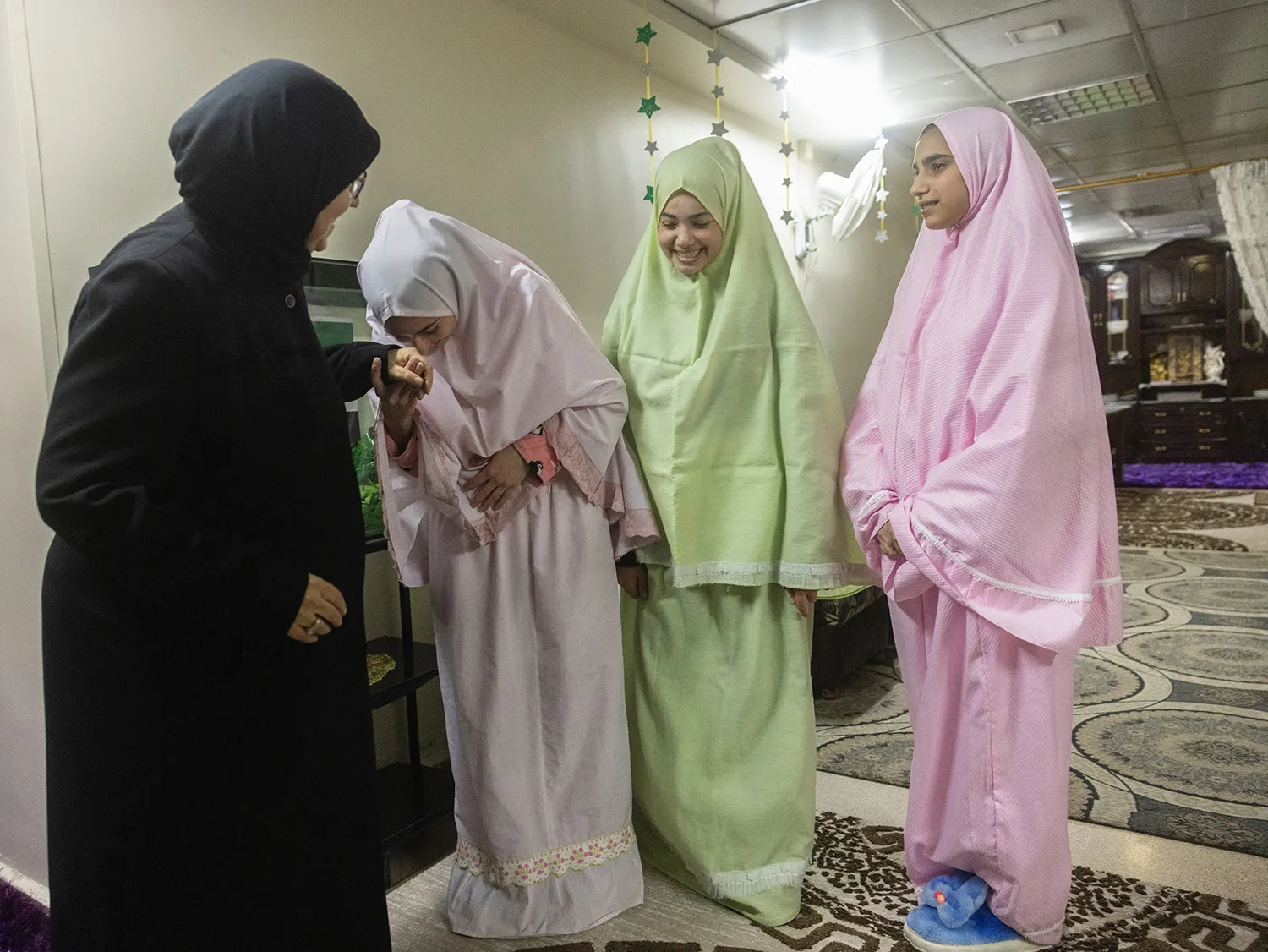 Three girls in pastel dresses and head coverings smile as they greet a woman in black headscarf and dress. One girl leans in to kiss the hand of the woman.