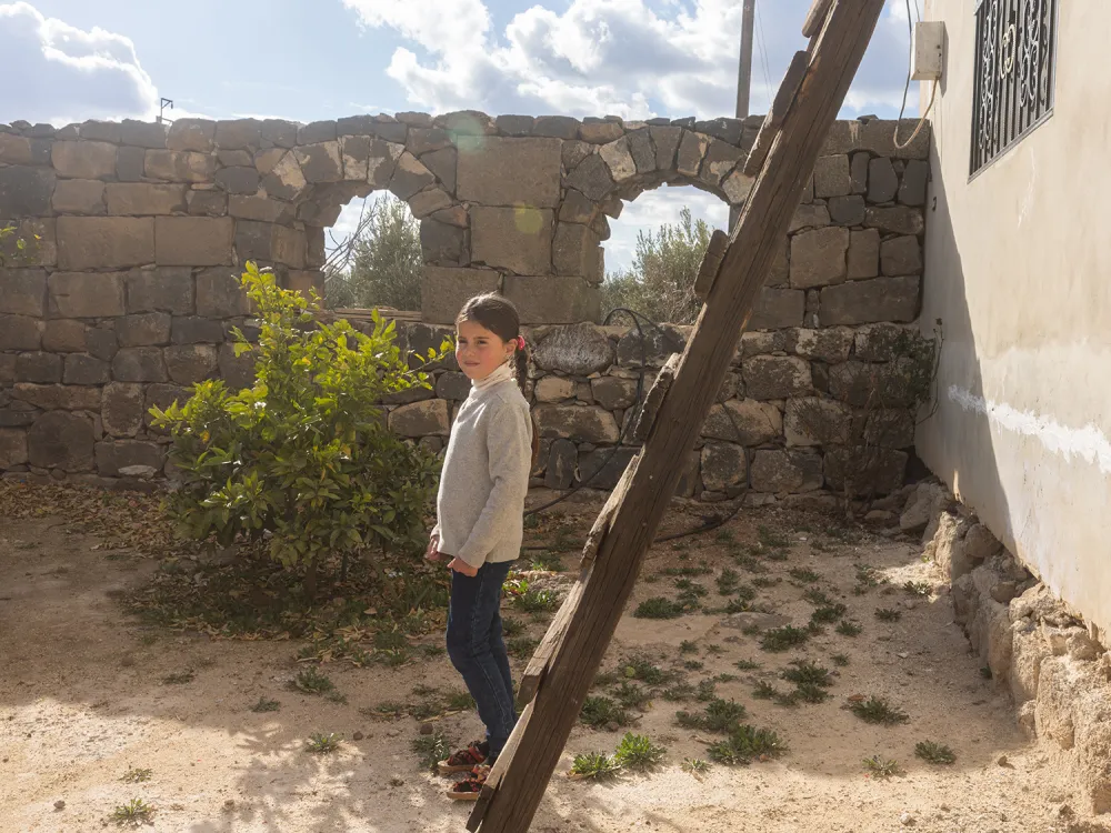 A girl of about 7 or 8 years old stands in front of a ladder propped against a wall in a yard with a more rough-hewn stone wall behind her, smiling slightly. Fluffy white clouds are visible in the bright sky overhead and through gaps in the stone wall.