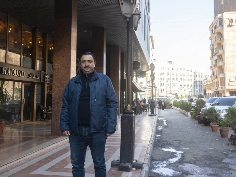 A man in a dark blue bomber jacket poses for a portrait on the sidewalk beside a damp street lined with midrise buildings on a sunny day.