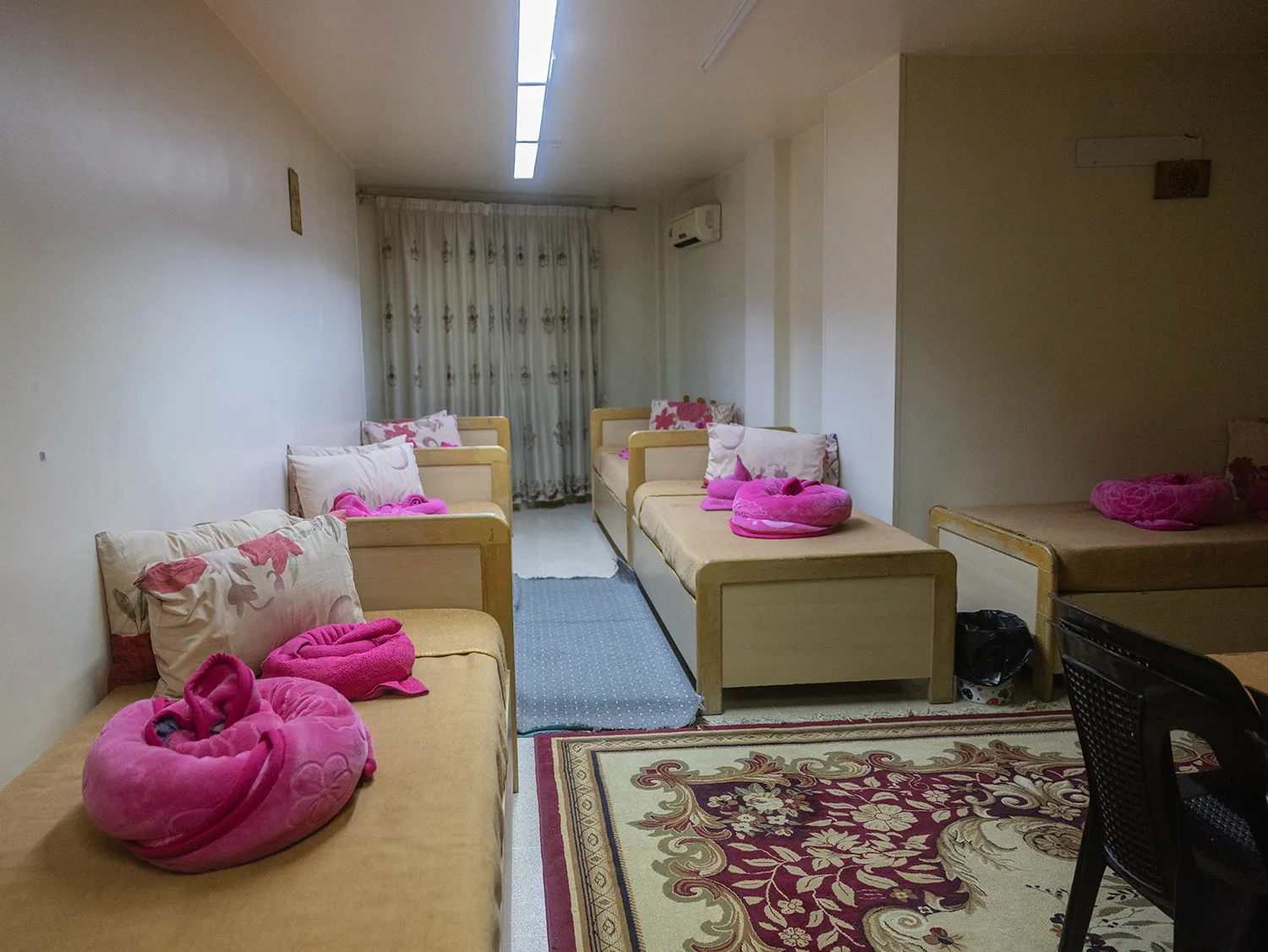 Three single beds stand in a narrow room with a floral rug and a long floral curtain stretched over the far wall. A desk is partly visible in the foreground.