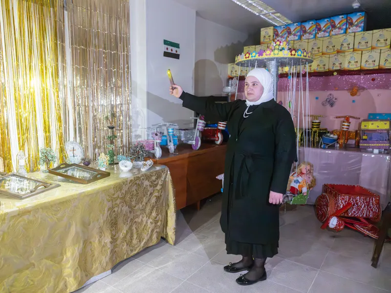 A woman in black with a white head covering uses her phone to light up a dim room that shows a tables covered with jewelry and craft items. Gold streamers hang over a window on the far wall.