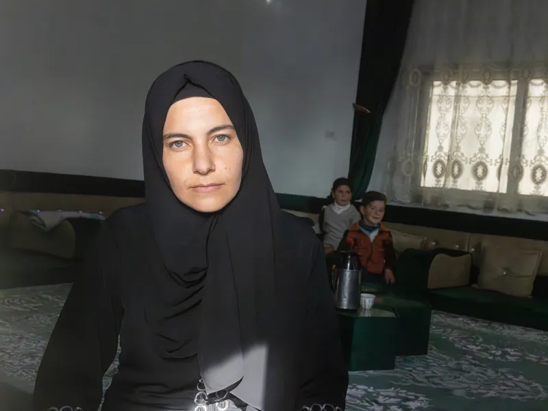 A gray-eyed woman wearing a headscarf and black dress sits in front of the camera. Behind her is a dimly lit room, where two young children perch on a chair, smiling as they eye the camera. The woman wears a serious expression and stares as the camera head-on.