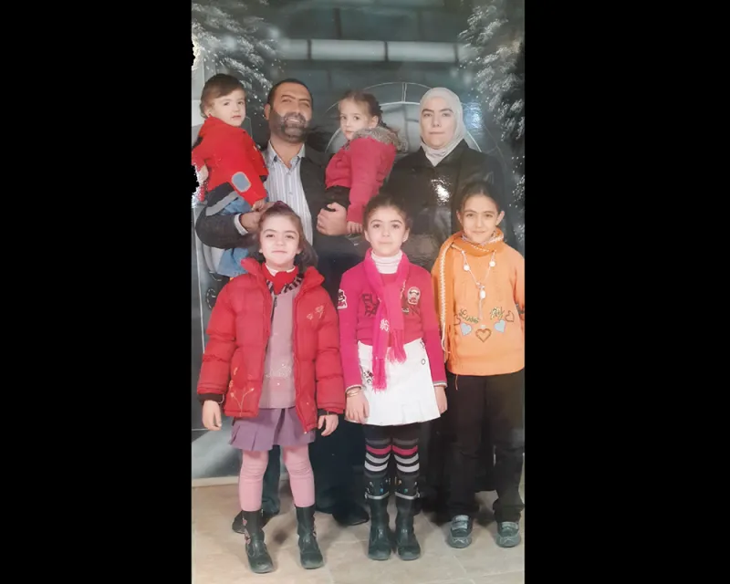 A man and woman and five children pose for a photo, most of them smiling for the camera. The family members are bundled in coats, scarves, and turtleneck shirts. The man holds two of the smallest children in his arms.