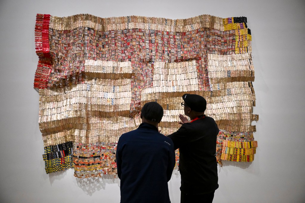 Two people view a large textile artwork made of small, colorful pieces.
