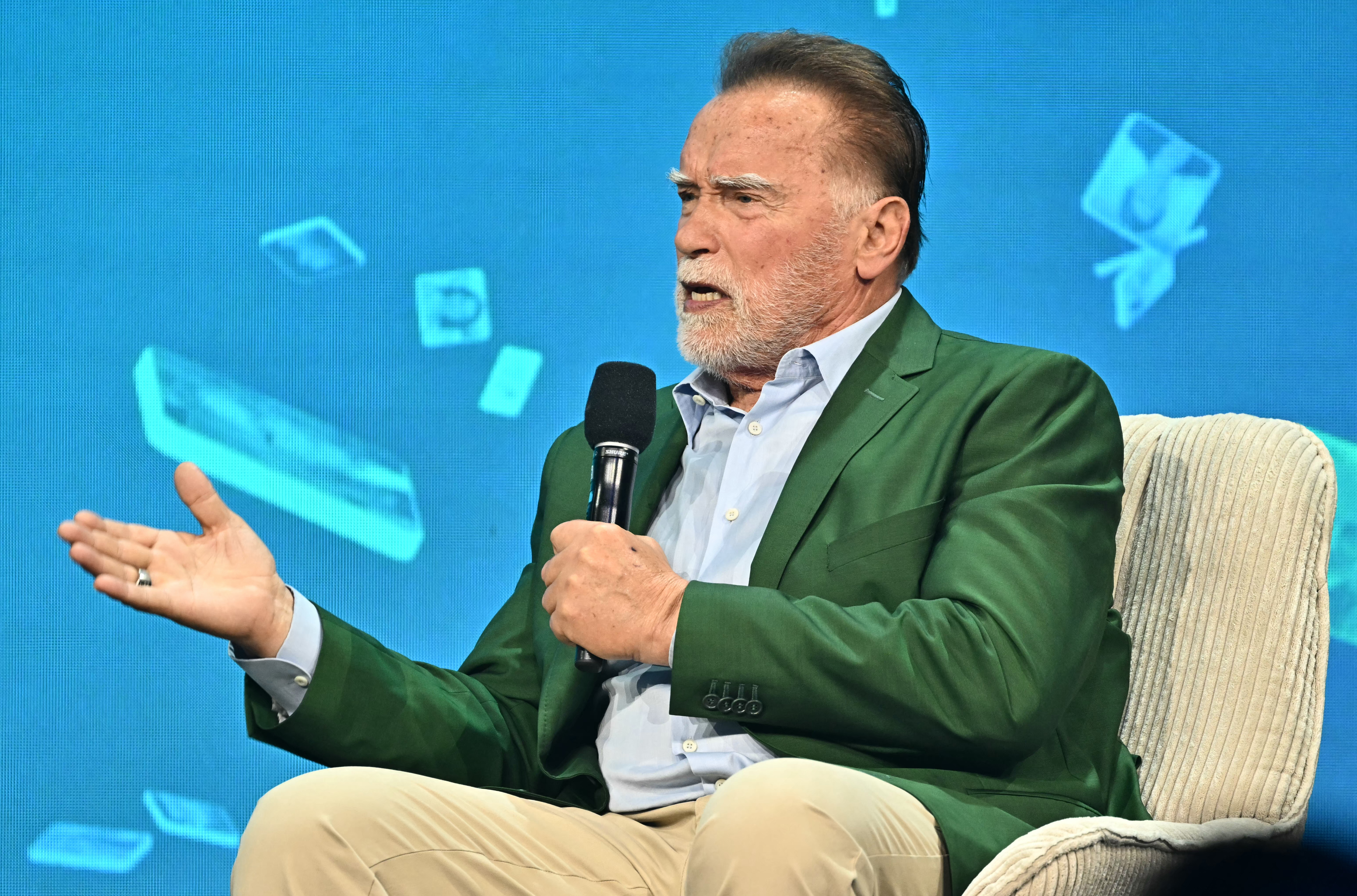 Arnold Schwarzenegger sits in a green jacket with a mic. 