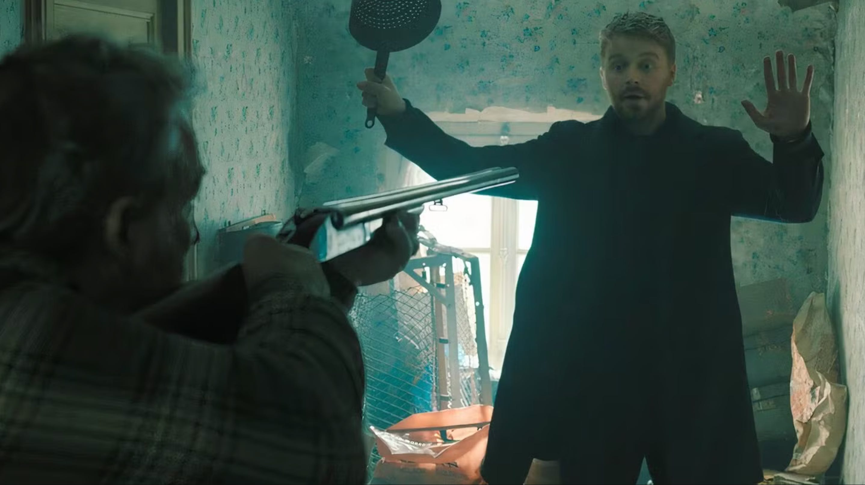 A man with light ginger hair and a beard in a long black coat, black sweater, and trousers holds his hands up in a ruined bedroom. He's holding a sieve in his right hand. An older man wearing a checked shirt in the left foreground is pointing a shot gun at the other man.
