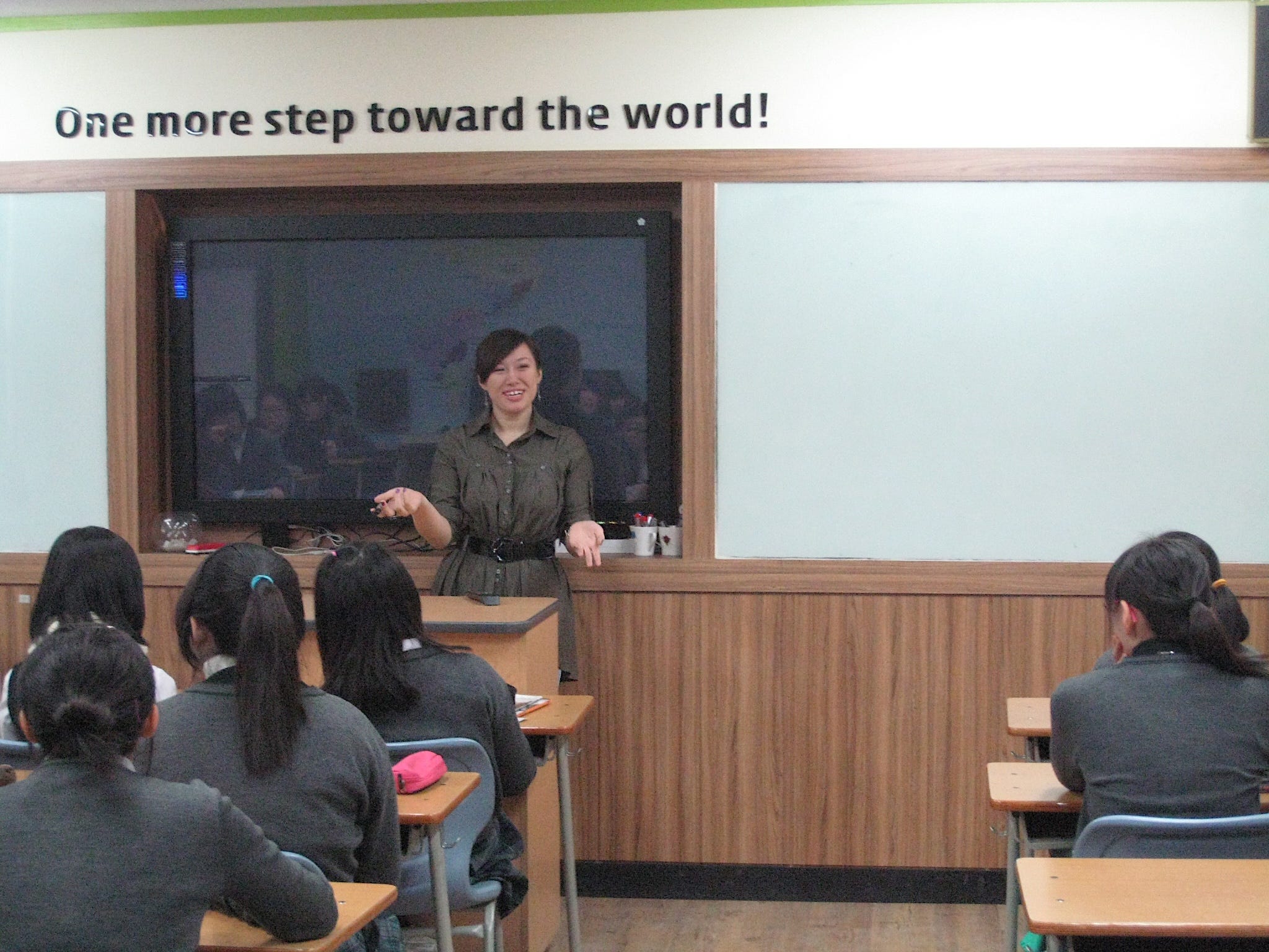 The author in the front of a classroom teaching a classroom of kids. The front of the classroom says "One more step toward the world!" above a screen.