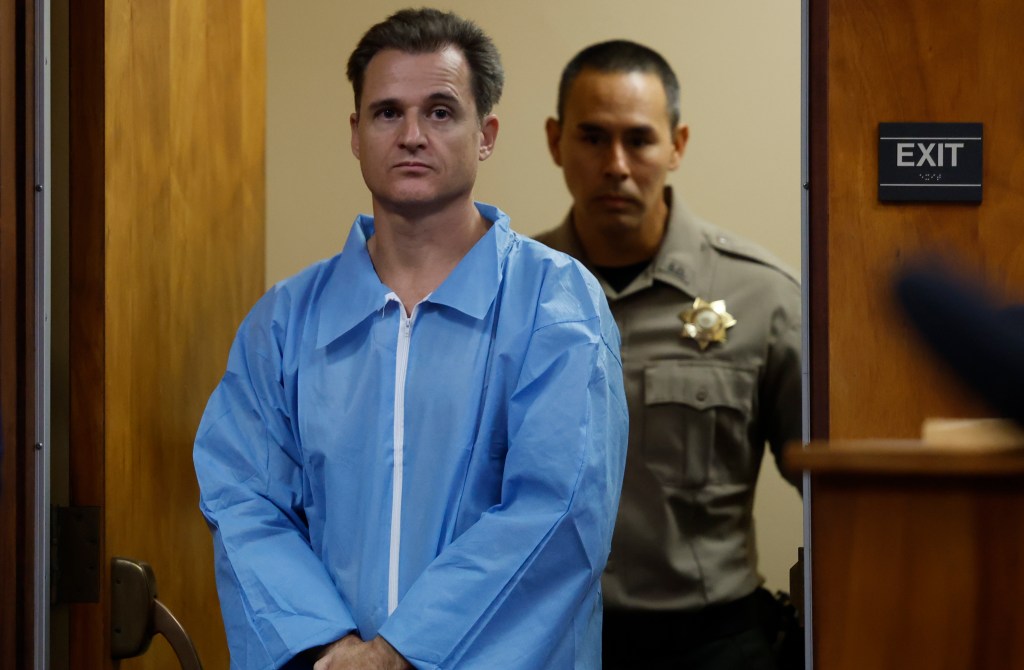 A doctor in a blue jumpsuit stands in a courtroom doorway, guarded by a uniformed officer.
