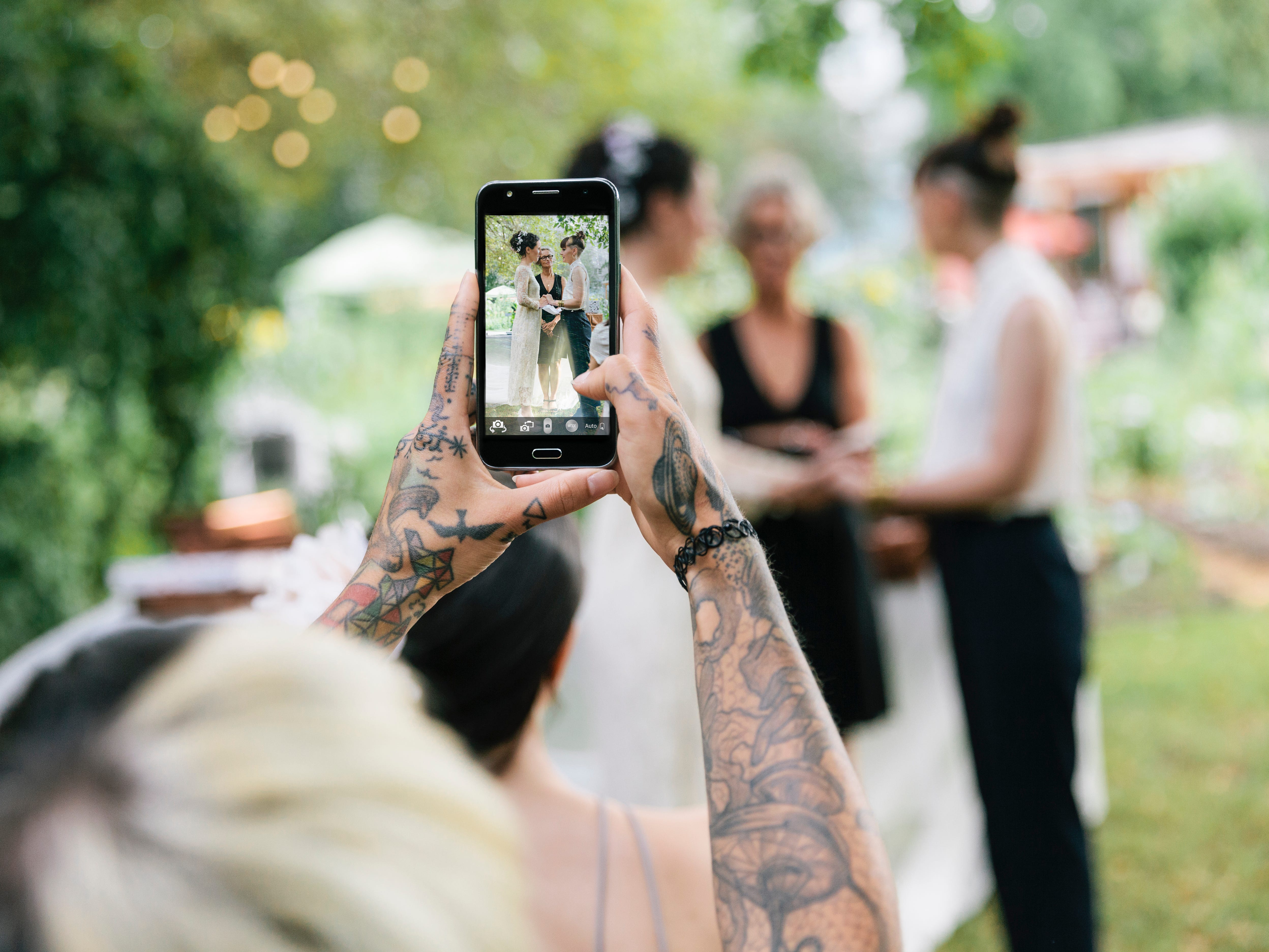 A tattooed woman holds up her phone to take photos of a couple during a wedding ceremony.