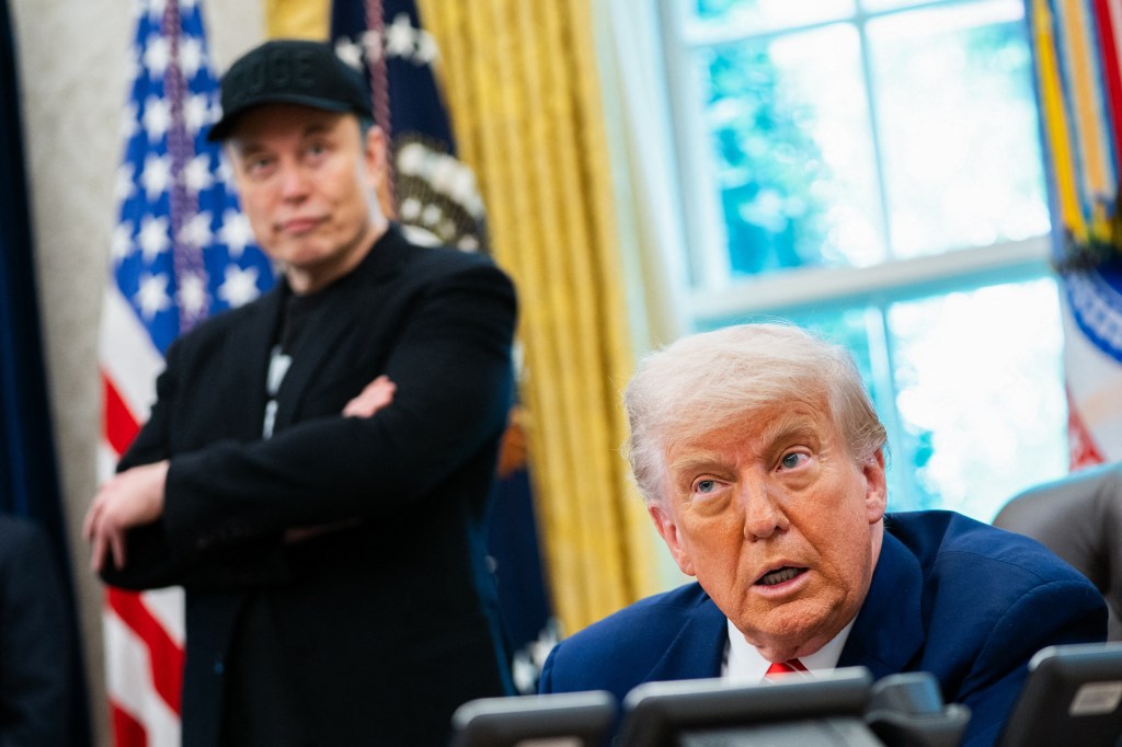 Donald Trump speaking at a press conference with Elon Musk standing behind him.