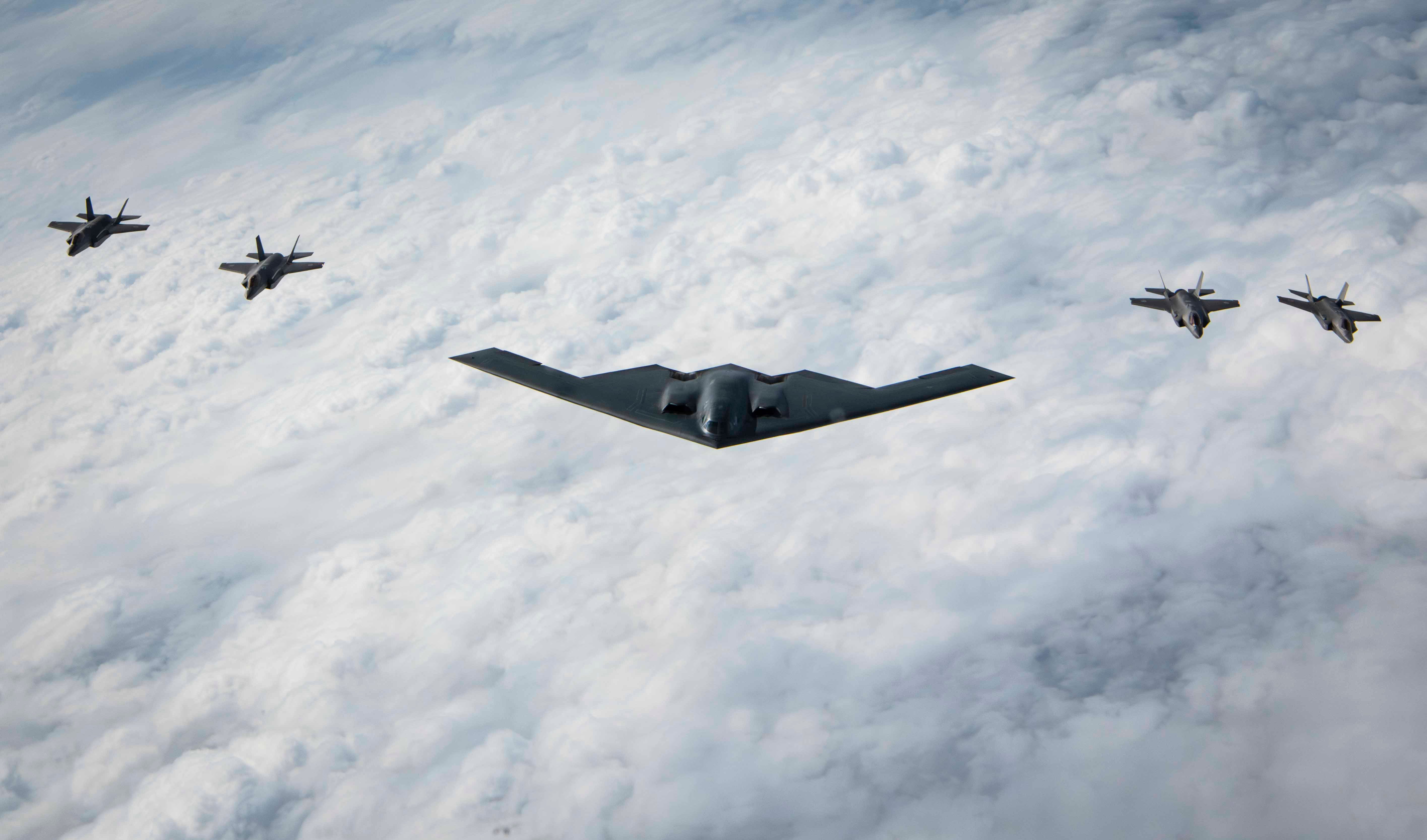 A B-2 Spirit stealth bomber flying above the clouds with two stealth fighter aircraft on either side of it.