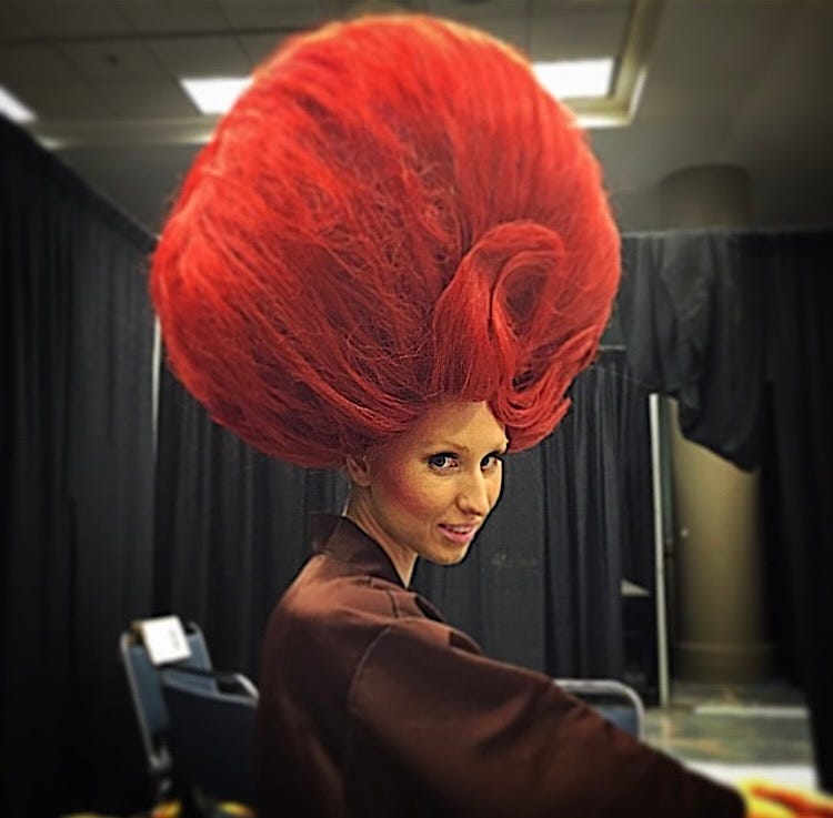 The author sitting in a chair getting her hair and makeup done. She has on a large red wig.