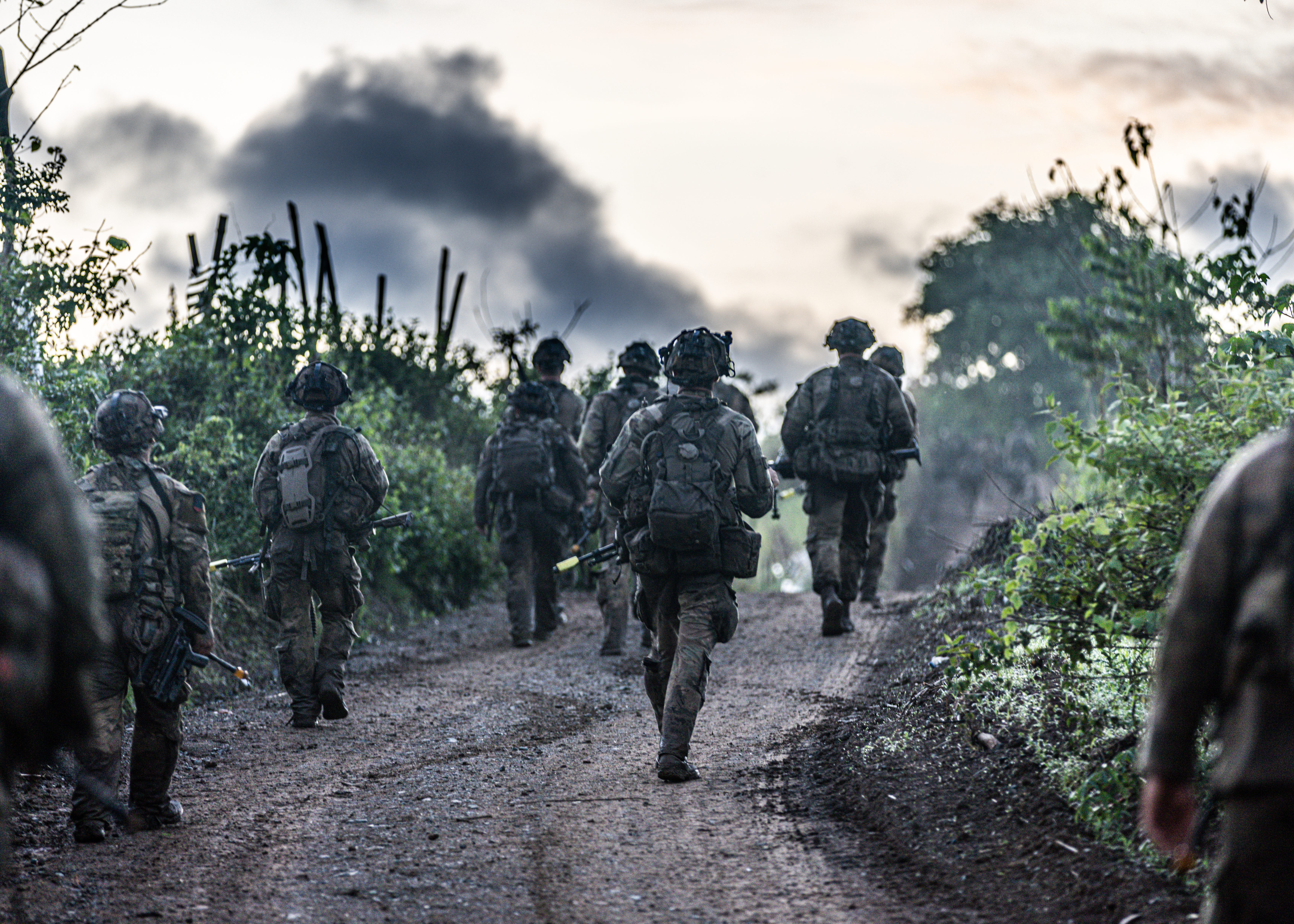 A group of soldiers wearing camouflage walk along a dirt road surrounded by green vegetation. Their backs are to the camera. The sky is cloudy and overcast.