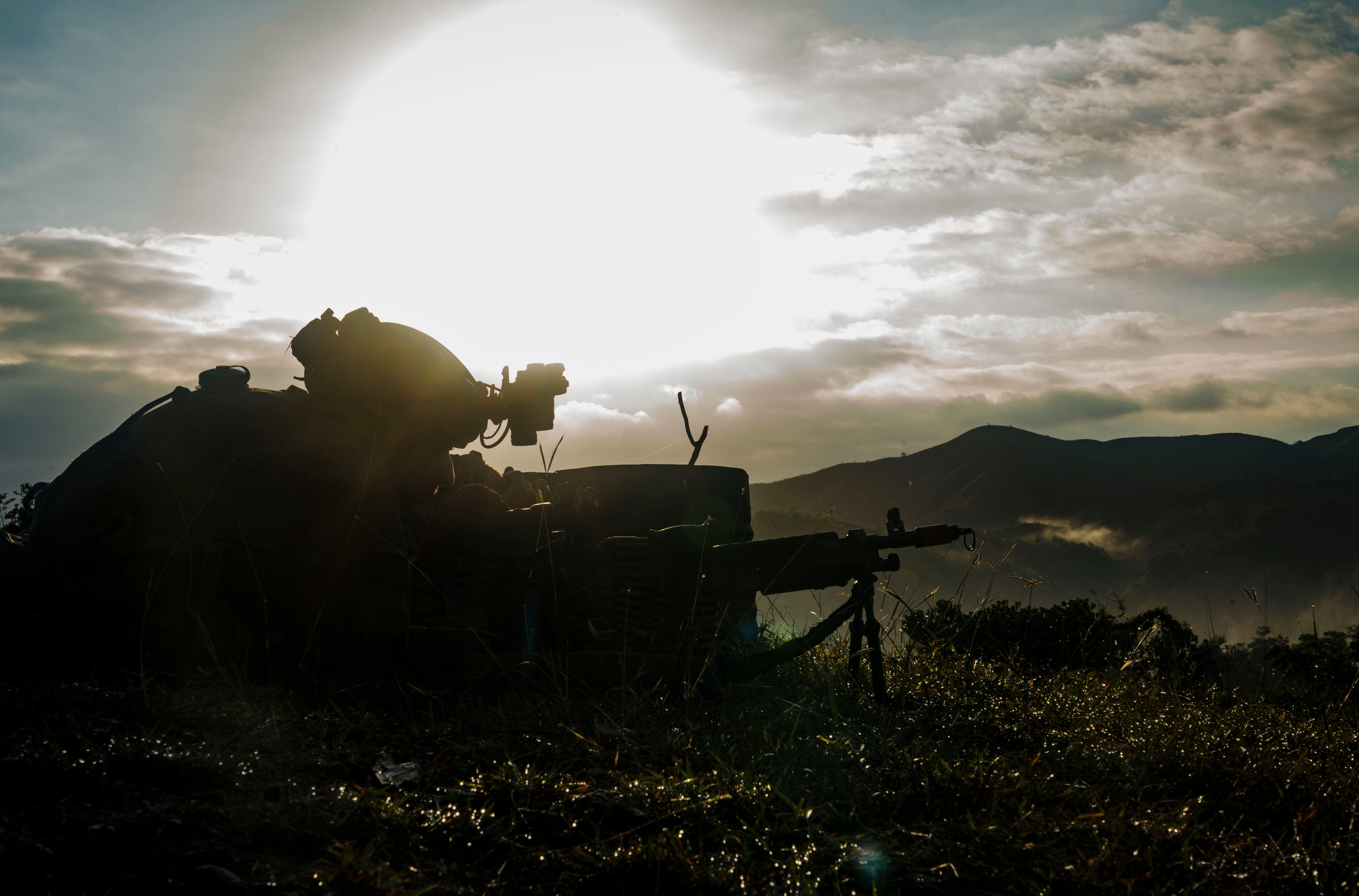 A side, shadowy profile of a soldier lying on the ground with a rifle. A mountain range and the sun in a cloudy sky are seen in the background.