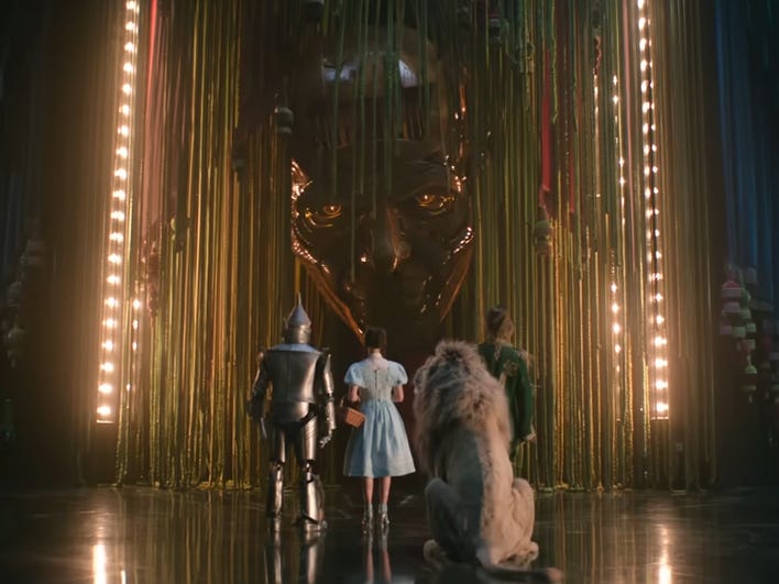 A screengrab of the "Wicked: For Good" trailer shows a Tin Man, a woman, a lion and a scarecrow standing facing toward a mechanical face.