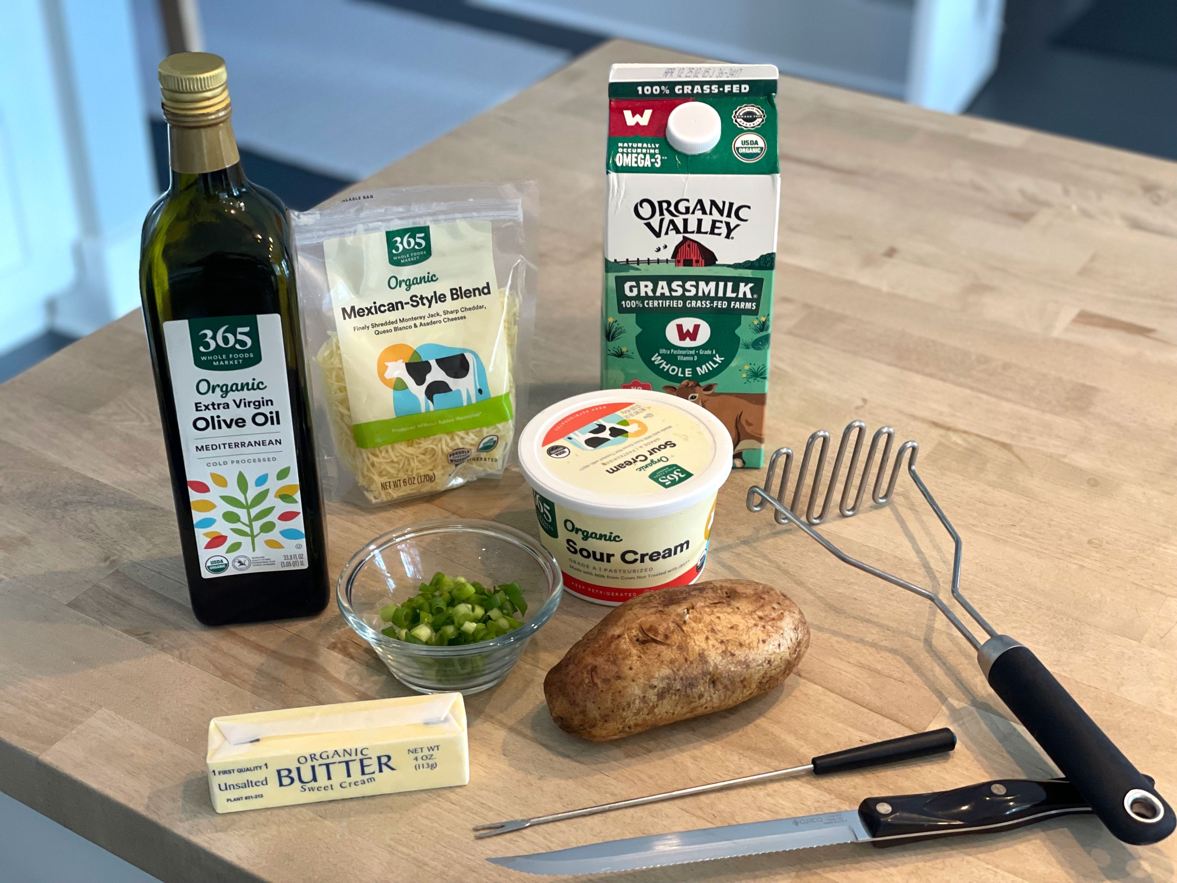 A bottle of olive oil, a package of shredded cheese, a carton of milk, a container of sour cream, a small bowl of green onions, a stick of butter, a potato, a knife, and other tools on a table.