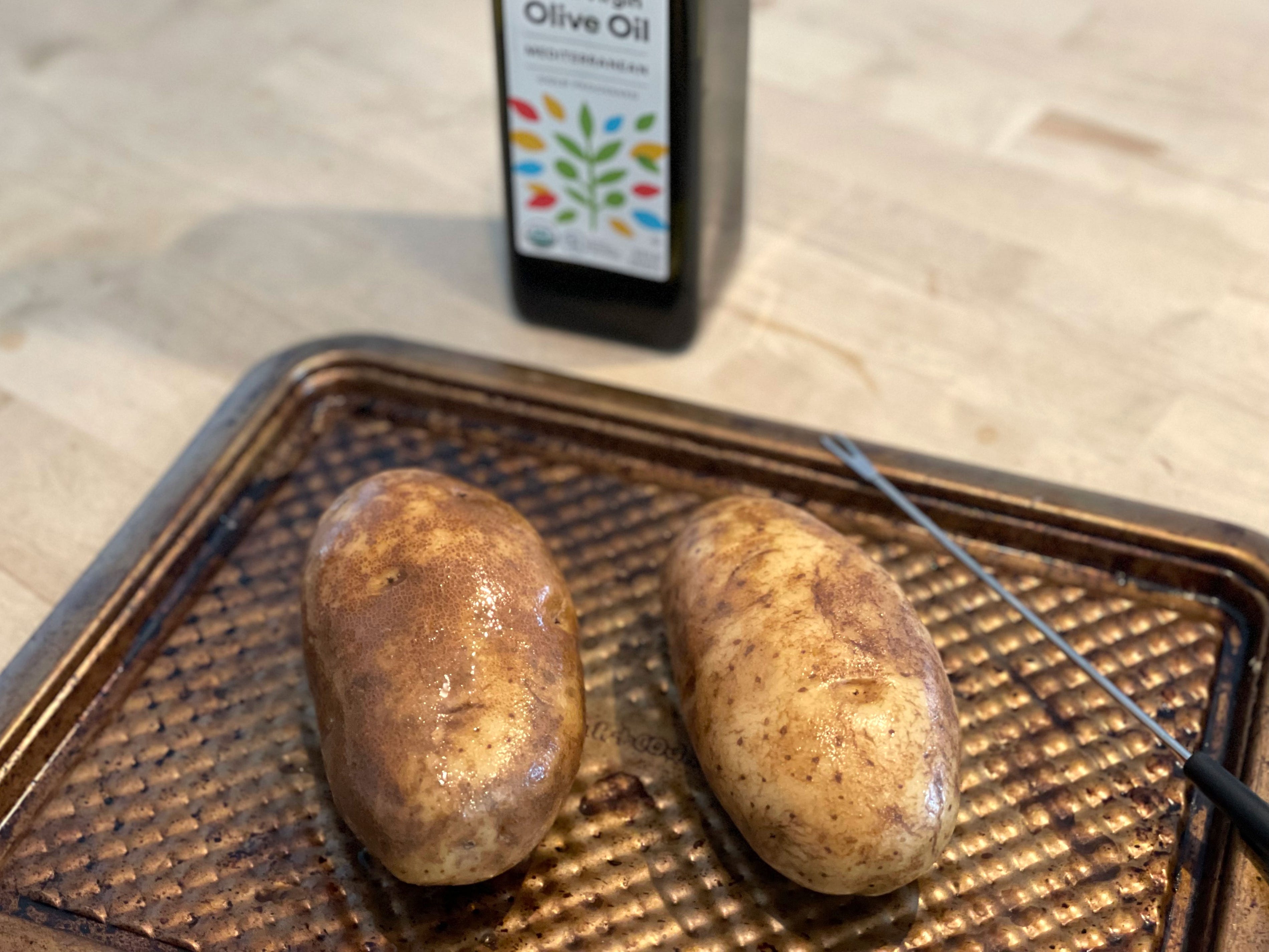 Two potatoes on a baking sheet with a bottle of olive oil in the background.