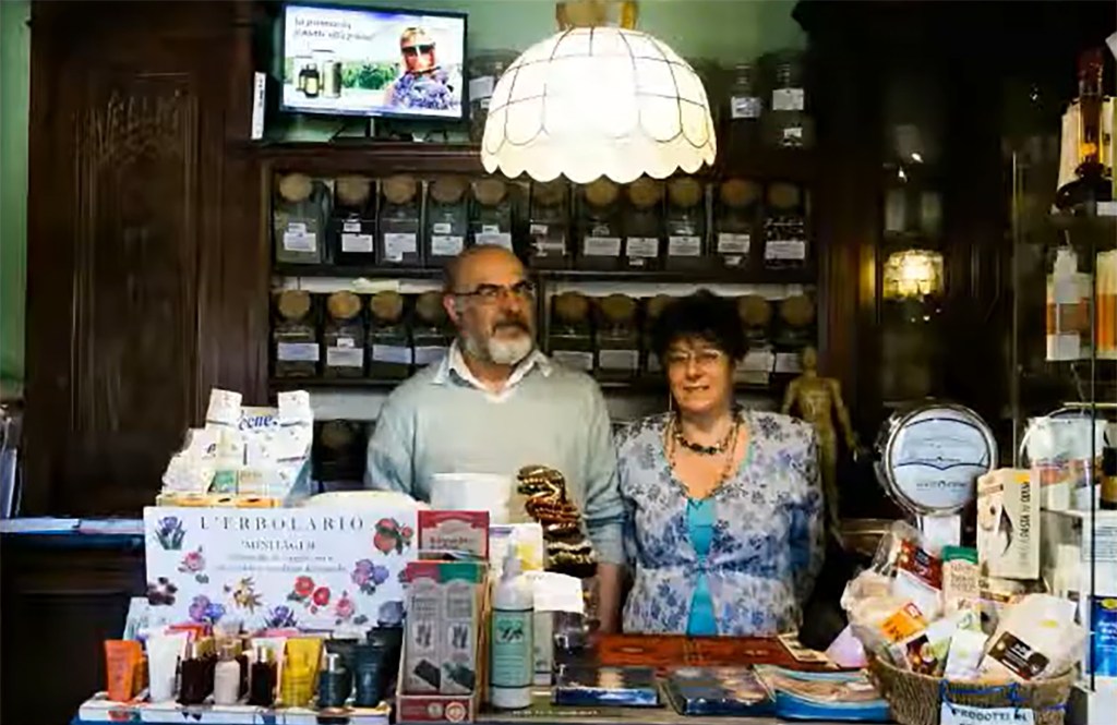 Maurizio and Lidia Carturan standing behind a counter in a shop filled with jars and boxes of herbs and other products.