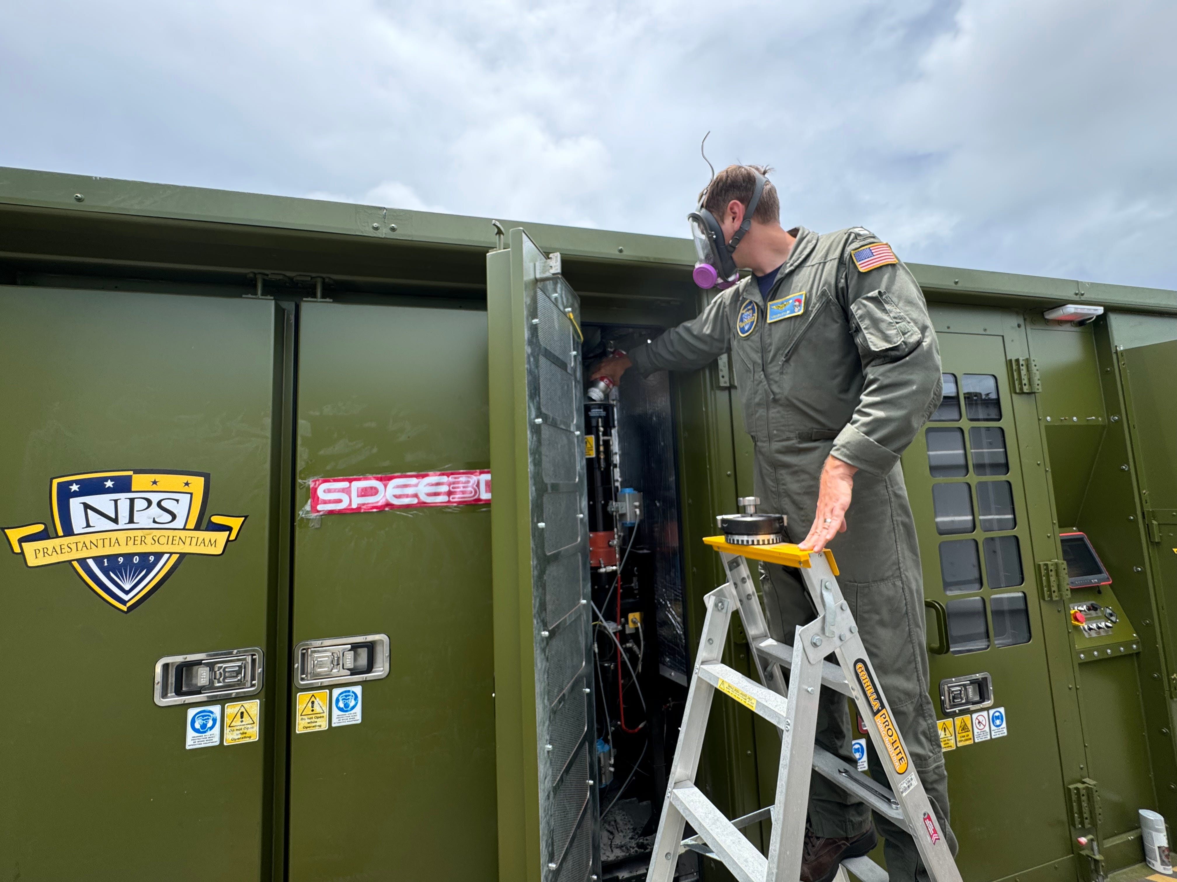 A man wearing a hunter green jumpsuit and a gas mask loads materials into a large Army green container. The sky is overcast above him.