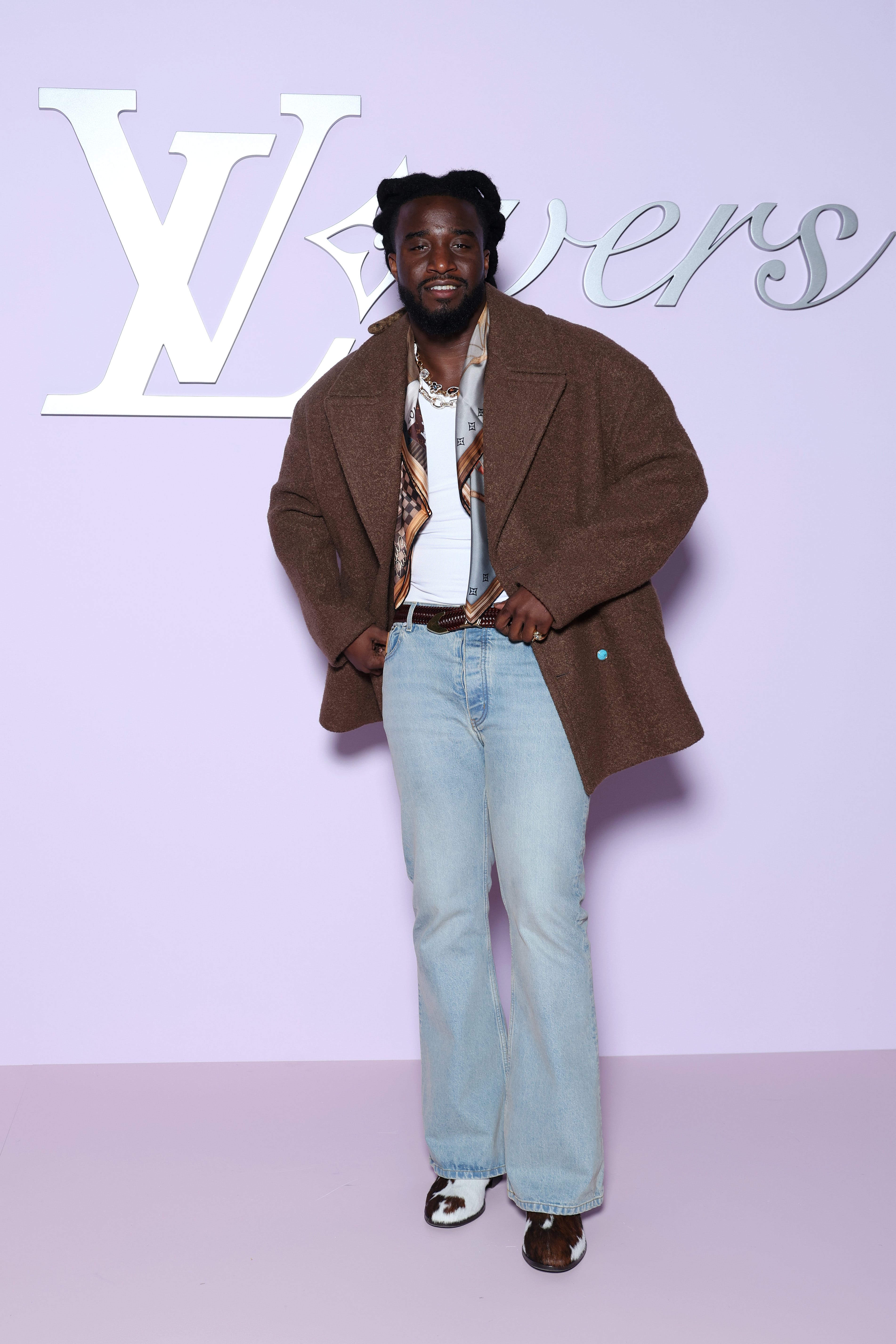 Shaboozey wears a brown jacket, white T-shirt, blue jeans, and a scar, at a Louis Vuitton runway show during Paris Fashion Week.