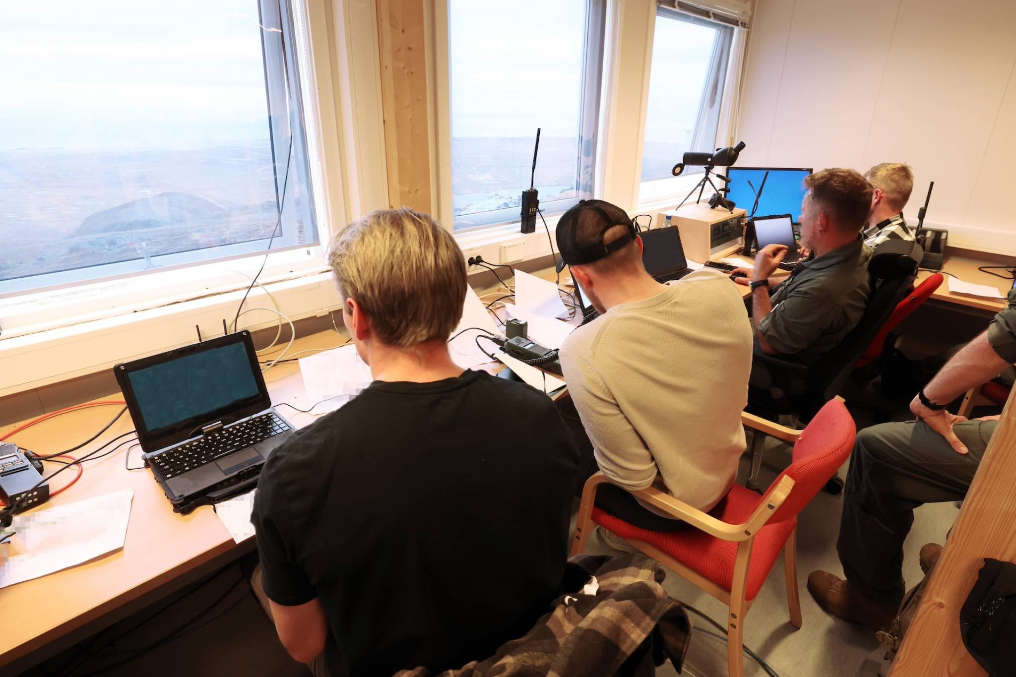 The backs of four men seated at a table against a window. Paper and computers are on the table.
