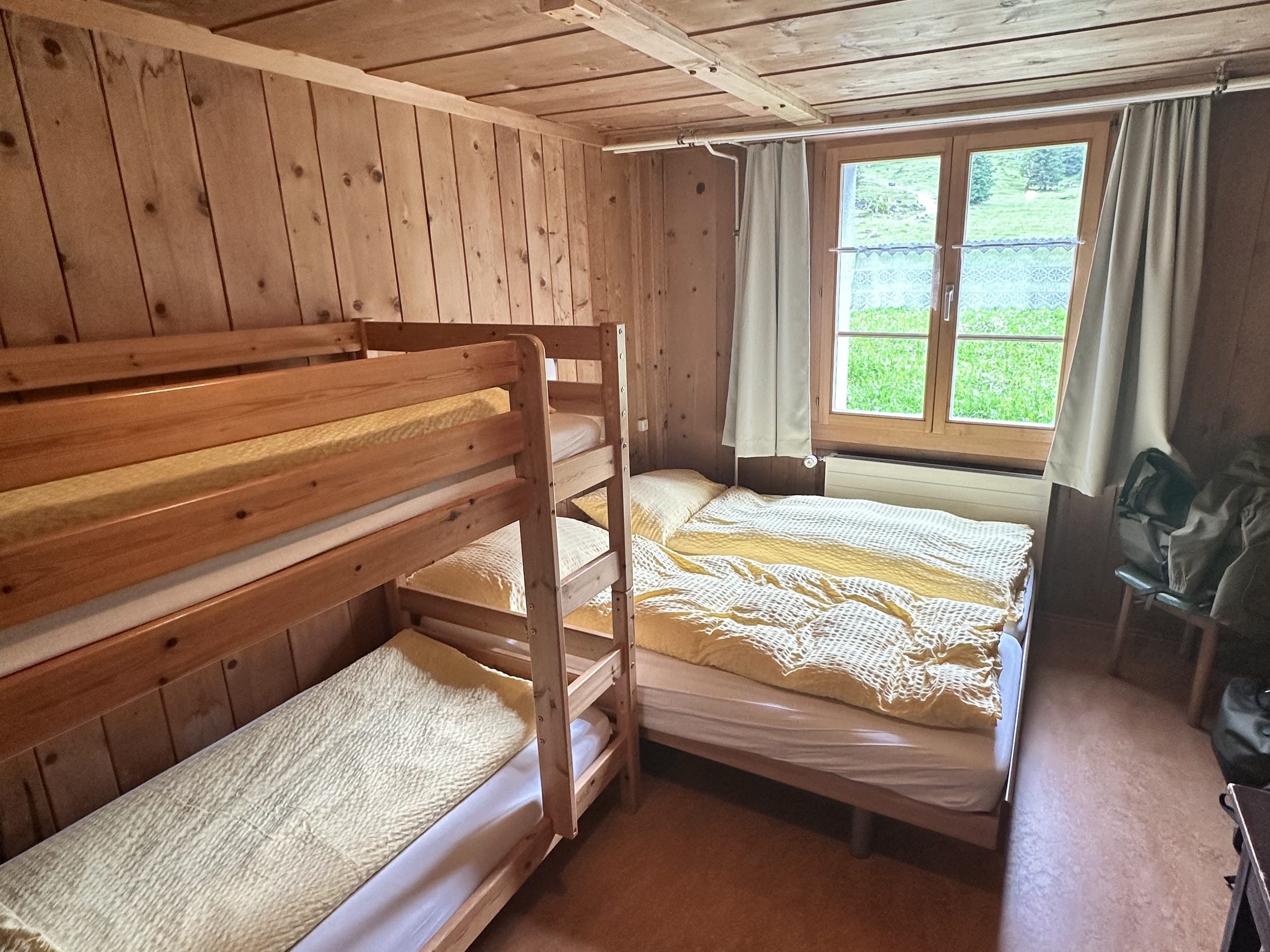 Inside a wooden cabin, a bed with two duvets next to a bunk bed and a window with green curtains showing a view of the grassy outdoors.