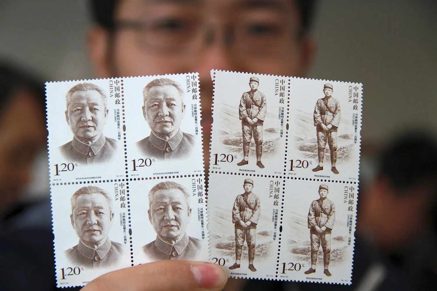 A man is seen out of focus as he holds up four each of two stamp designs depicting Xi Zhongxun. One design shows a close-up portrait of his head; the other design shows him standing in a military uniform in front of a hilly landscape