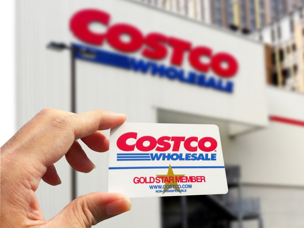 A hand holding a Costco Gold Star membership card with a blurred Costco warehouse in the background.