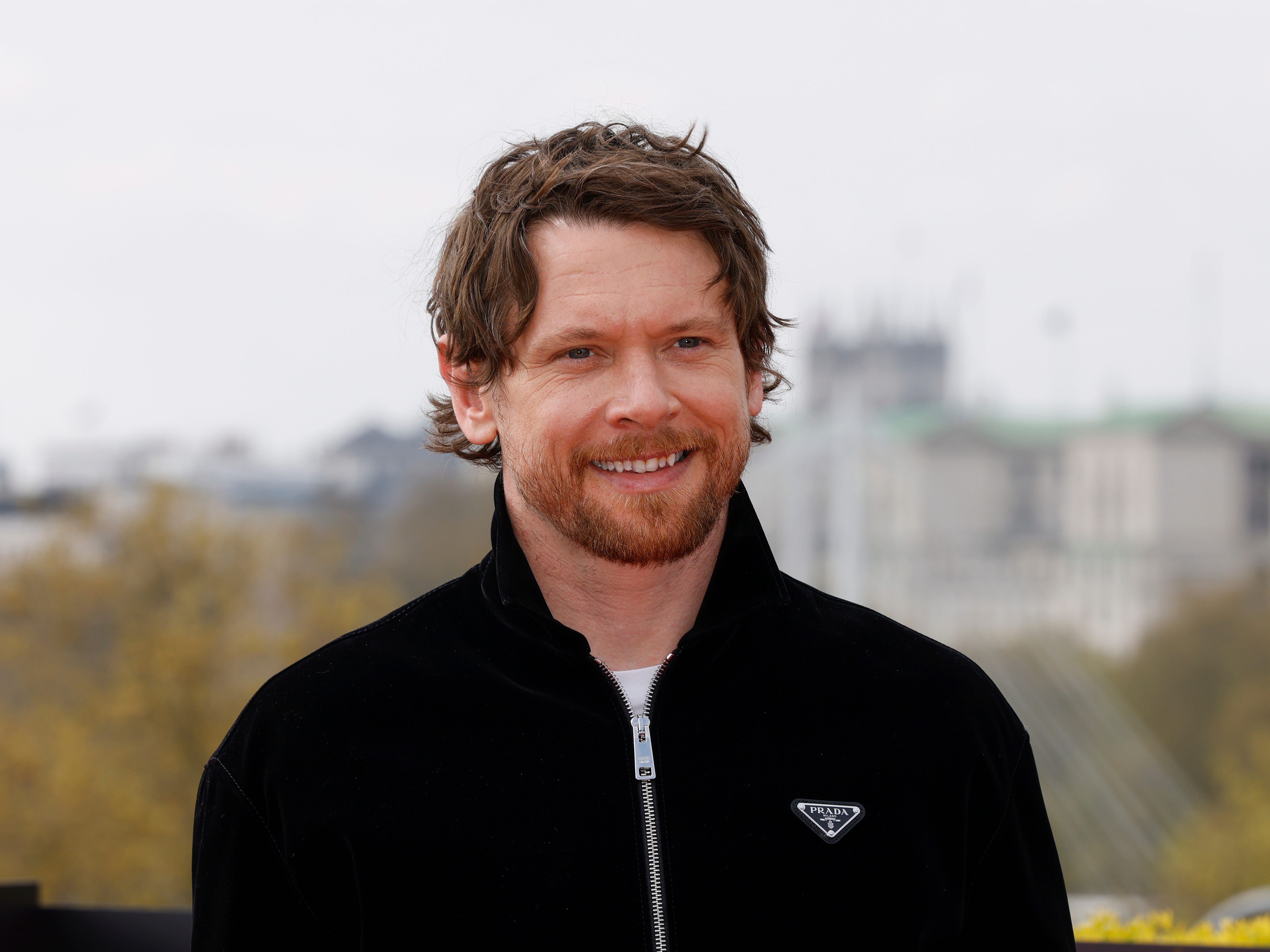 A man with messy brown hair stands with a blurred city in the background. He's got a brown beard and is wearing a black zipped-up jacket with a triangular 