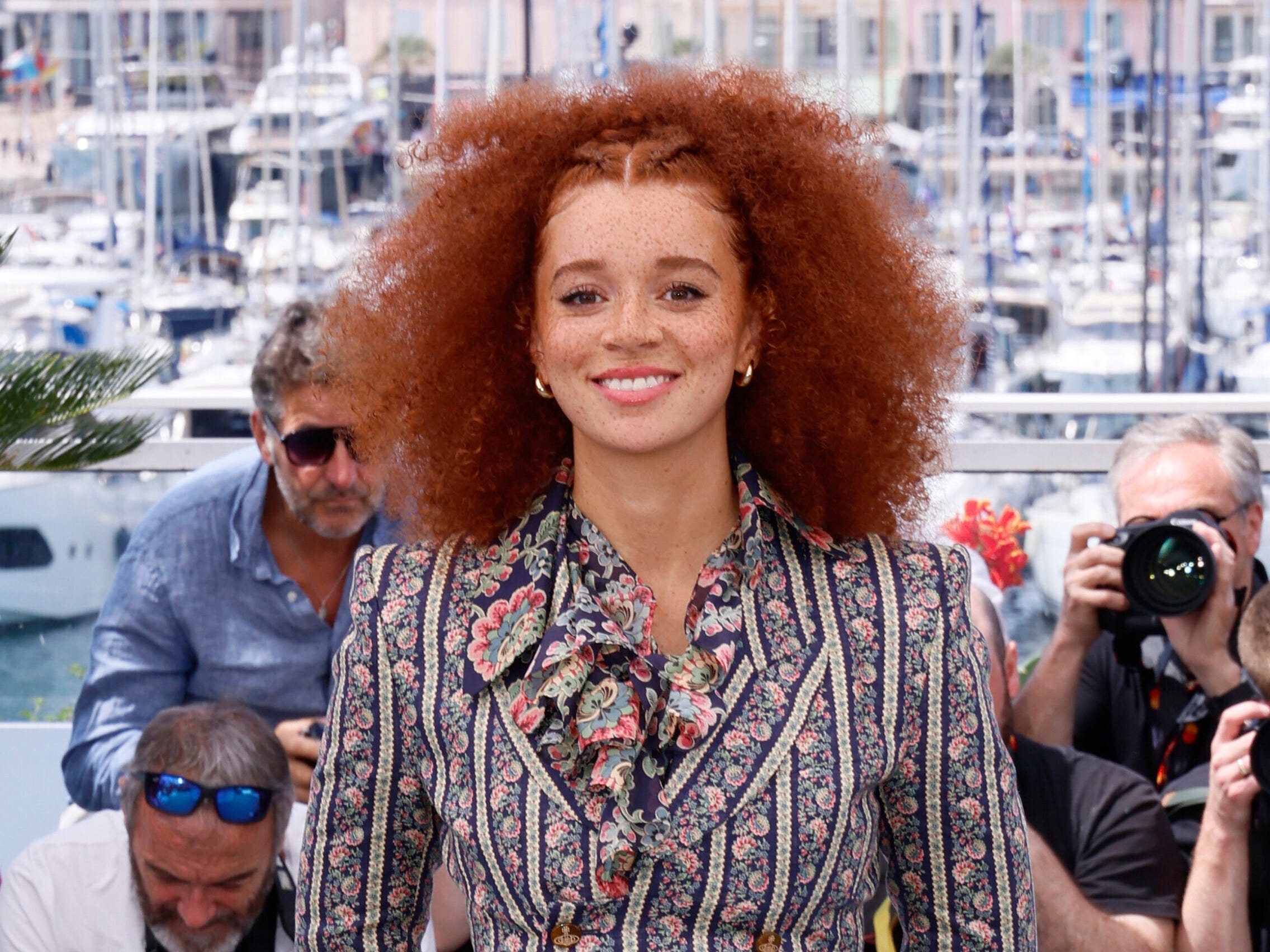 A woman with frizzy ginger hair smiles for the camera. She's wearing a floral striped suit with a floral, frilly shirt underneath the blazer.